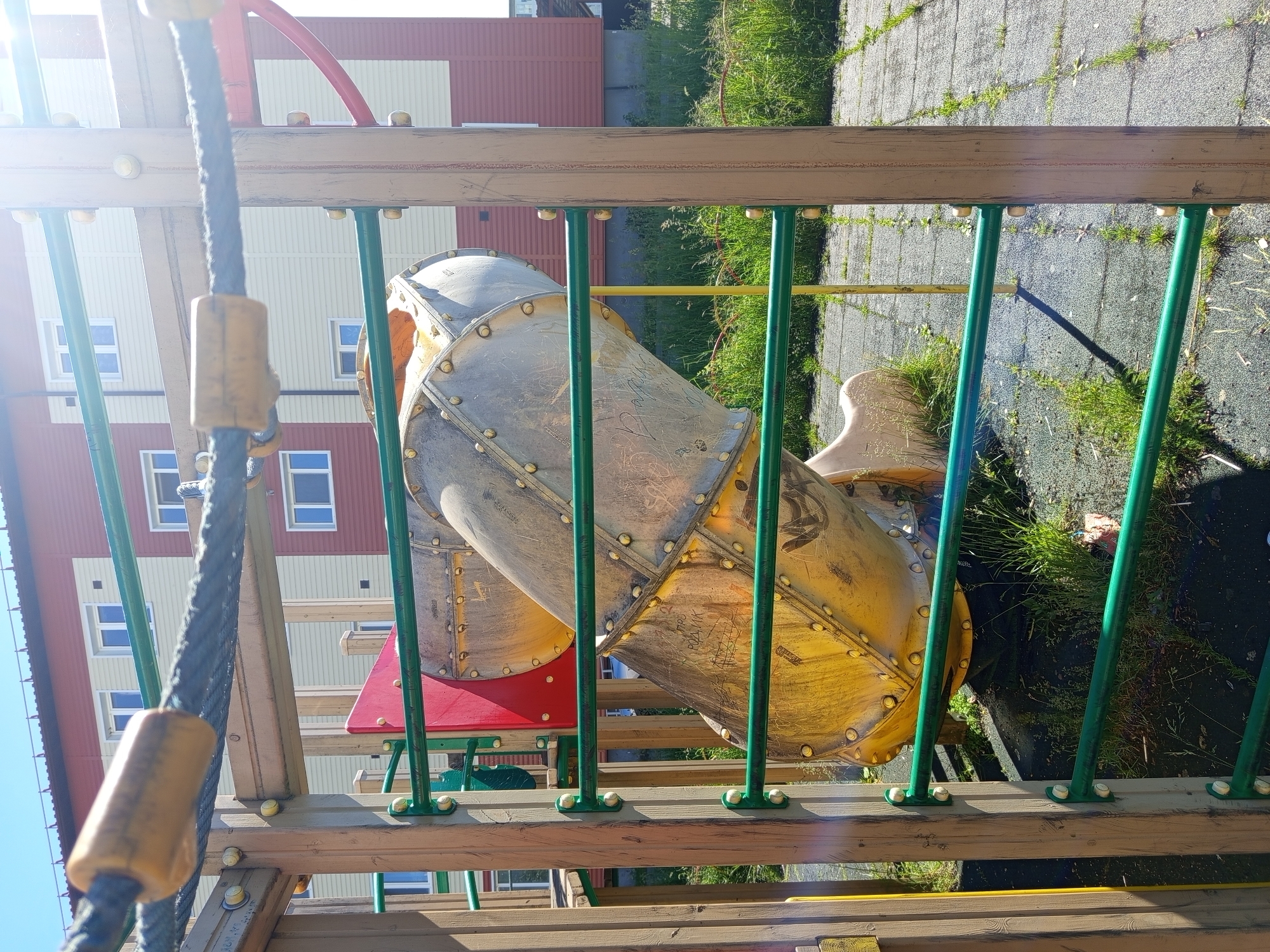 The image depicts a playground slide structure, made of yellow and silver metal, with green horizontal bars in the foreground. A dark rope with yellow cylindrical attachments is stretched across the top of the frame. In the background, a modern apartment building with red and cream-colored siding and white-framed windows is visible. The ground is covered with gray rubberized tiles, interspersed with grass and weeds. The scene suggests an outdoor setting in an urban or suburban area. This scene is located in the city of Gdov, Russia.