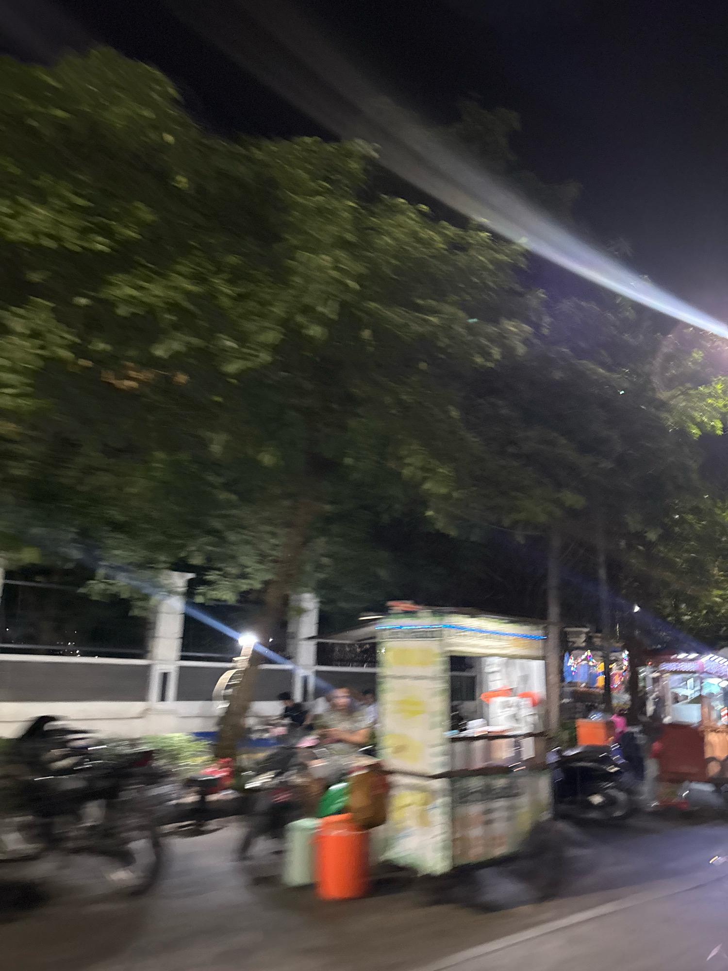 A night-time scene in North Jakarta, Indonesia, captures a street with several food stalls and parked motorcycles. The image is blurred due to motion, likely from a moving vehicle. Large, leafy trees are prominent, their foliage appearing dark against the night sky, illuminated by a bright light beam from the right, possibly a spotlight or from a street lamp.
The foreground shows the blurry outlines of several motorcycles parked along a paved road. To the right of the motorcycles, there are food stalls set up. One stall in the center has a colorful, illuminated facade and appears to be selling food. There are also orange and green bins visible near the stalls.
In the background, a white wall with rectangular windows stretches along the side of the road. Several people are present, but their features are indistinct due to the motion blur and low light conditions. One person is visible sitting at a table near the stall.
The overall impression is of a lively street scene at night, possibly a market or a gathering spot, with the motion blur adding a dynamic, fleeting quality to the captured moment.