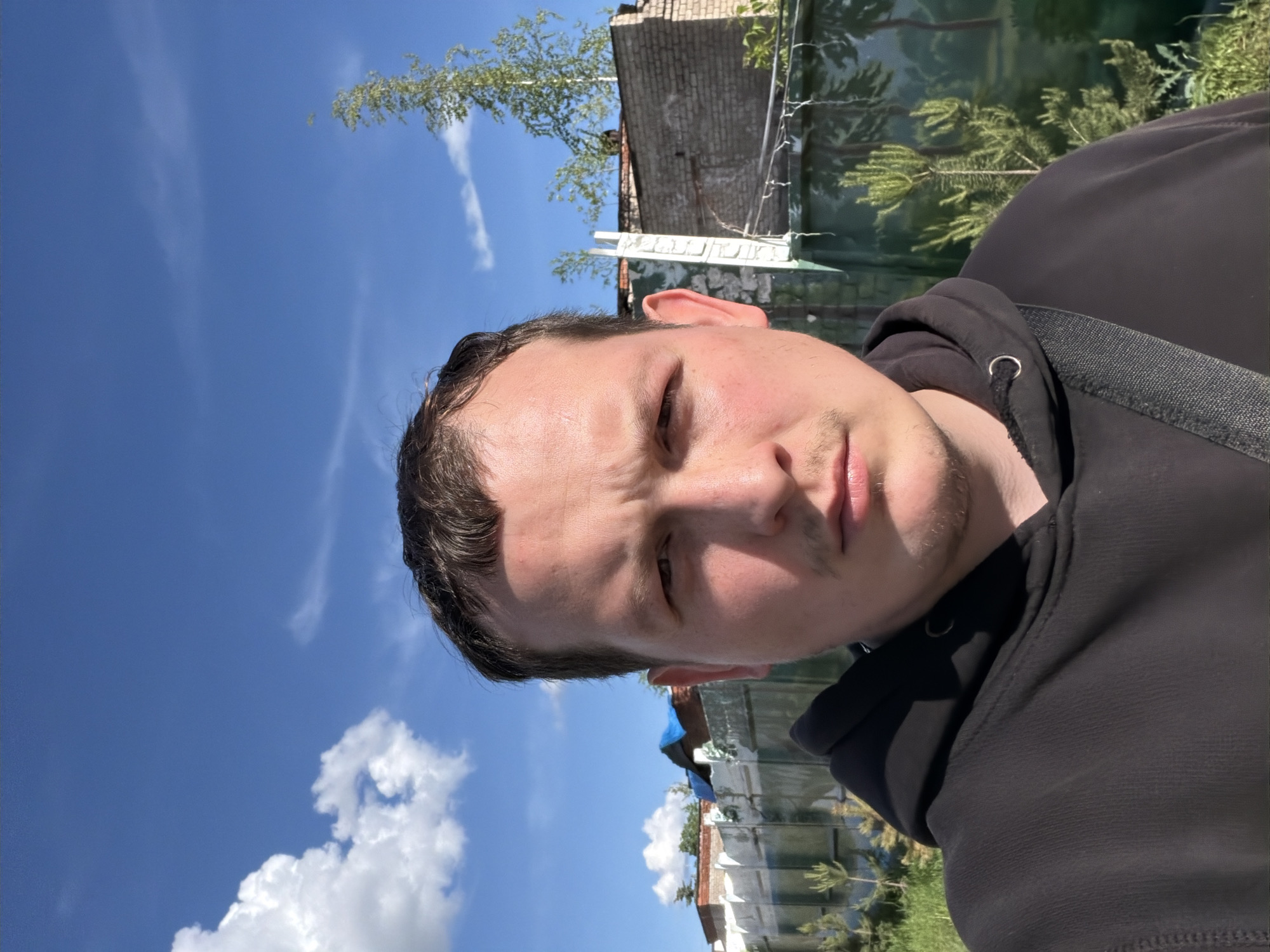 A man is taking a selfie outdoors under a blue sky with white clouds. He is wearing a black hoodie and a strap is visible across his body. In the background, there are buildings and trees. The location appears to be in a rural or suburban area. The exact location within the city of Chaikovsky, Russia is not discernible from the image.