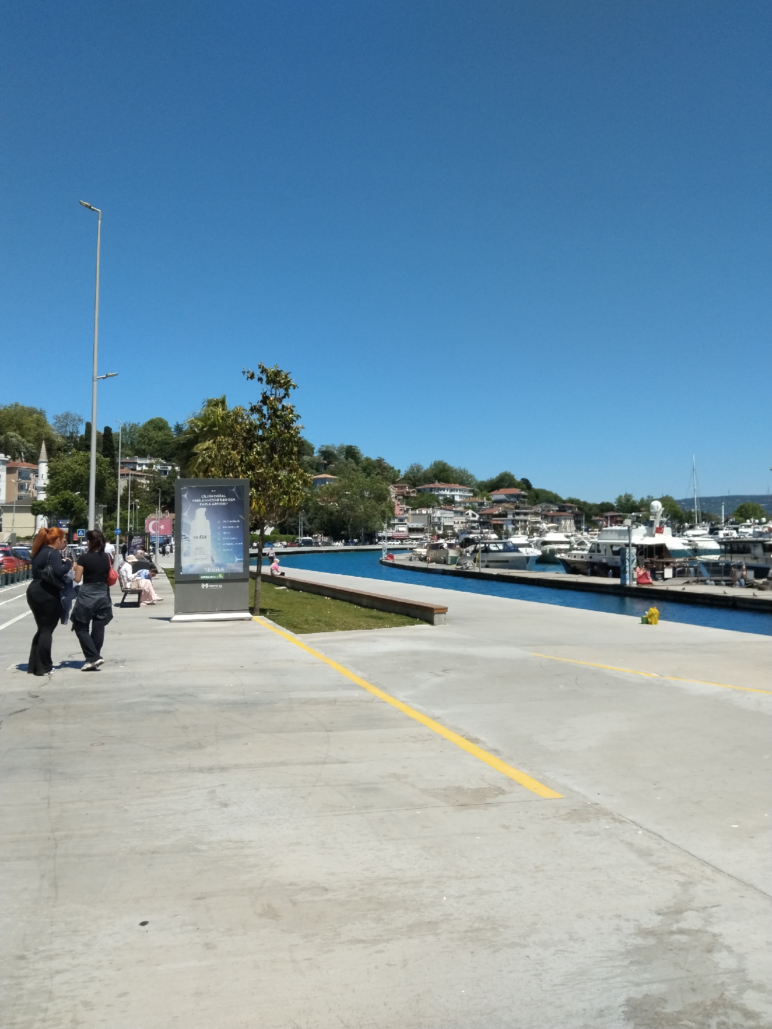 The image captures a bright, sunny day at what appears to be a marina. The sky is a clear, deep blue. A concrete pedestrian walkway is visible in the foreground, marked with a yellow line. To the left, two women are walking along the walkway. Further down the walkway are benches where people are sitting. A digital advertising kiosk stands beside a small tree. Beyond the walkway, a narrow strip of green grass separates it from a canal filled with boats. Various sizes and types of boats are docked along the canal. Buildings and trees rise on the opposite bank of the canal.