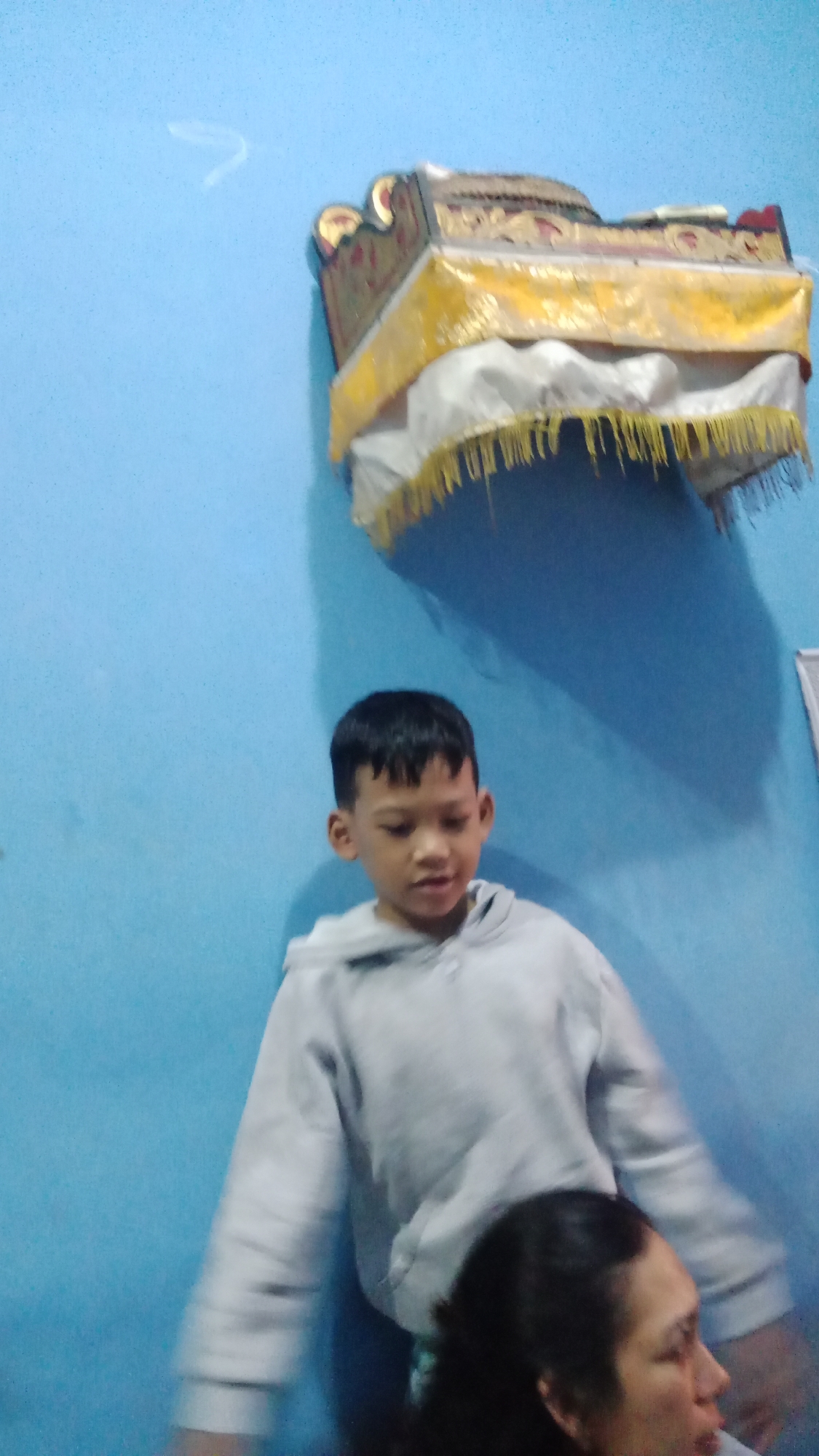 In an indoor setting with a light blue wall, a young boy wearing a light grey hooded sweatshirt stands and looks downwards. Above and to the right of the boy, a decorative, ornate object is mounted on the wall. This object appears to be a ceremonial altar or shrine, with gold and red decorative elements and white and yellow fabric draping from it. In the lower right corner of the image, a portion of an adult's head and shoulders is visible, with the individual facing to the right and out of frame. The image quality is blurry, particularly the lower portion depicting the adult. This scene is consistent with a domestic interior in Denpasar, Indonesia, likely a home or a place of worship.