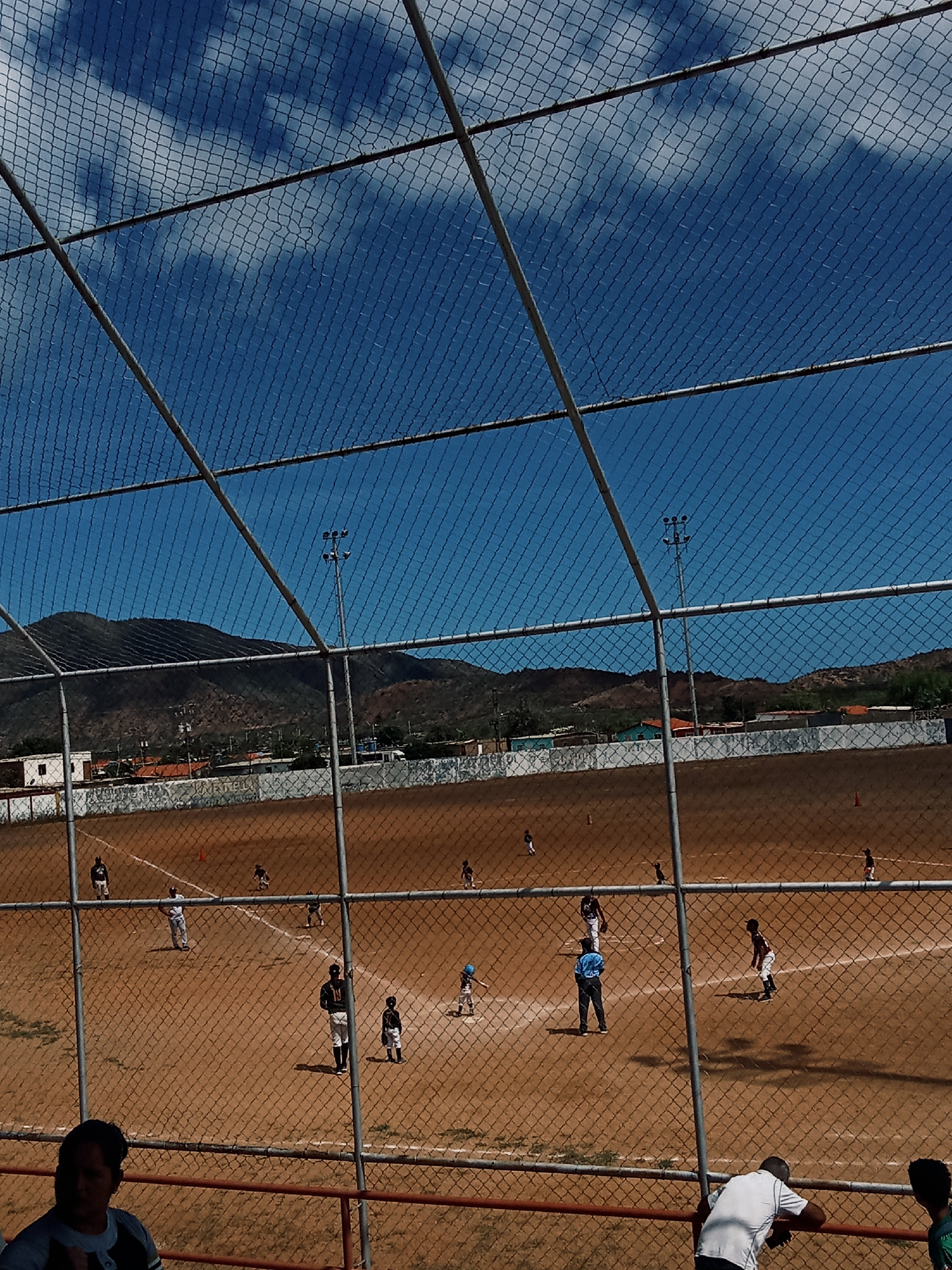 A baseball game is in progress at a baseball field in Juan Griego, Venezuela. The view is from behind a chain-link fence, partially obscuring the field. The field is brown dirt with white base lines and cones. Players in black and white uniforms are scattered across the field, some in motion and some stationary. A man in a blue shirt is near the center of the field. There are buildings along the edge of the field and mountains in the background, under a blue sky with white clouds. In the foreground, people are visible behind a metal railing.
