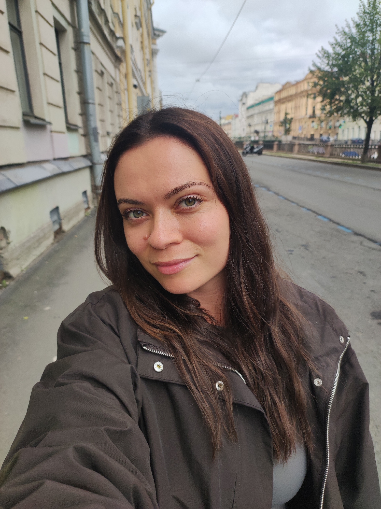 A medium shot selfie shows a woman with long, dark brown hair and green eyes. She is wearing a dark brown jacket over a light gray top. The woman is standing on a sidewalk next to a street with a tree-lined embankment in the background. The architecture in the background suggests the location is Saint Petersburg, Russia, likely on a canal embankment, possibly the Griboyedov Canal, given the style of buildings and the presence of a tree. The sky is overcast.