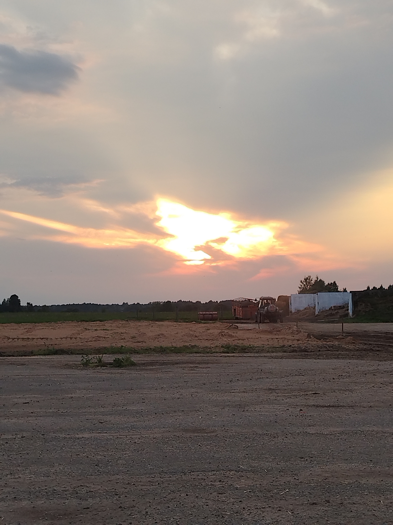 The image shows an outdoor scene at dusk or dawn, likely in a rural or agricultural area. A tractor is visible in the middle ground, positioned near a white rectangular structure. To the left of the tractor, there are some industrial containers and what appears to be stacked hay or straw. The foreground consists of a dirt or gravel road. In the background, there is a treeline and a cultivated field. The sky is dramatic, with the sun breaking through clouds and casting rays of light, creating a luminous effect. The overall impression is one of a working agricultural landscape at the end or beginning of the day. The image has been tagged with the location of Завуццеўскі сельскі Савет, Belarus.