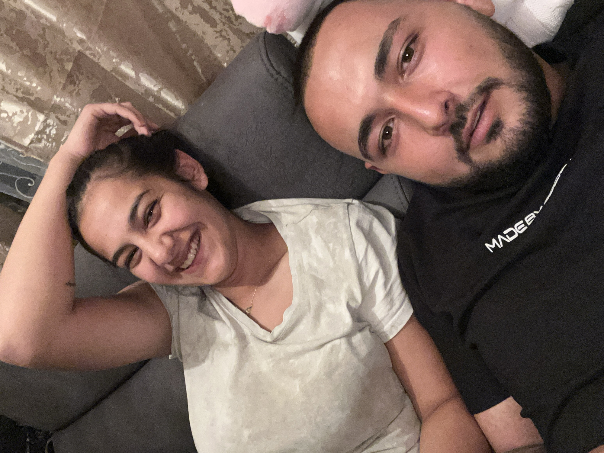 The image depicts two individuals lying on a gray upholstered surface, potentially a sofa or a bed. A woman on the left is smiling and has her left hand touching her hair. She is wearing a light-colored t-shirt and has a necklace. A man on the right is closer to the camera, with a beard and a black t-shirt with "MADE" visible. The background includes curtains with a patterned design. The location in Razhevo Konare, Bulgaria, is unknown, as there are no location specific features in the image.