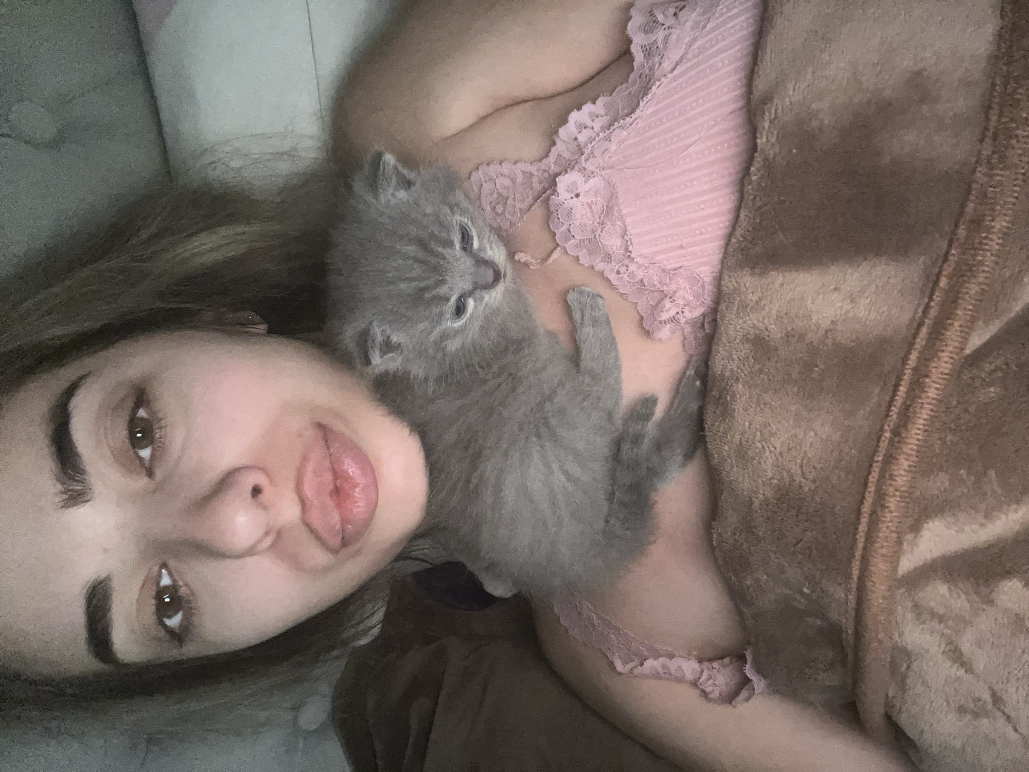 A close-up selfie shows a woman holding a small grey kitten in her arms. The woman, whose face is partially visible, has dark hair and is wearing a pink lacy top. The kitten is nestled against her chest. The background consists of a soft, brown blanket and a light-colored surface, possibly a pillow or bed. The image appears to have been taken indoors, in a bedroom setting. There is no visible indication of the location being in Velykyi Bychkiv, Ukraine.