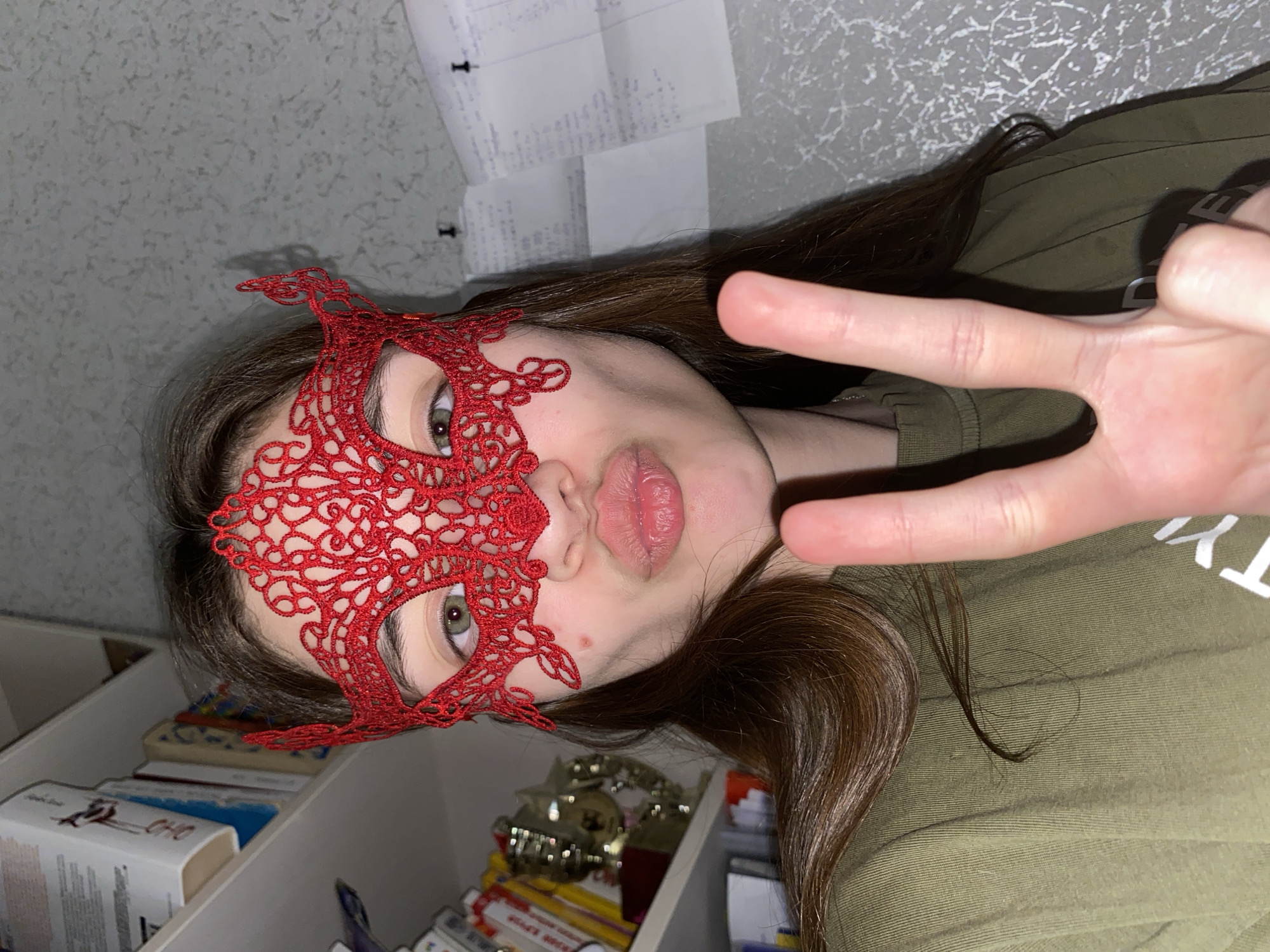 An indoor, portrait-style photograph captures a young person in Votkinsk, Russia, positioned at a diagonal angle. The subject, with long brown hair, wears a vibrant red lace mask covering their eyes and forehead, through which their green eyes are visible. Their lips are slightly pouted, and a small blemish is visible on their right cheek. They are dressed in a green or khaki long-sleeved shirt, with partial white lettering, possibly "TY", visible on the right side. The person's right hand is raised, forming a peace sign (V-sign) towards the camera.
The background suggests a personal room or study. To the left, white shelves hold books and what appear to be small, shiny decorative items or trophies. Faint text like "ОНО" and "CHO" is partially visible on some book spines. Above and behind the subject, a light-colored, possibly textured wall is visible, to which several sheets of paper containing indistinct printed text are affixed with small, dark pins. The lighting is consistent with an indoor setting, possibly under artificial light. The overall impression is one of playful posing.