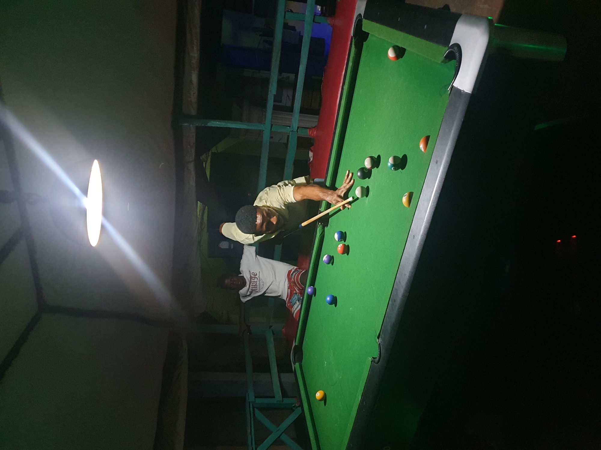 An evening shot shows two men playing pool in Lokoja, Nigeria. The men are positioned on opposite sides of a green pool table, illuminated by a single hanging light fixture. One man, wearing a light-colored t-shirt and patterned shorts, is seated with his arms outstretched, watching the game. The other man, dressed in a light yellow shirt, is actively engaged, poised to strike a billiard ball with a cue. Several pool balls are scattered across the table, some in play and some already pocketed. The background reveals a dimly lit, open-air structure with green wooden railings, suggesting a casual, recreational setting. The overall atmosphere is relaxed and focused on the game.