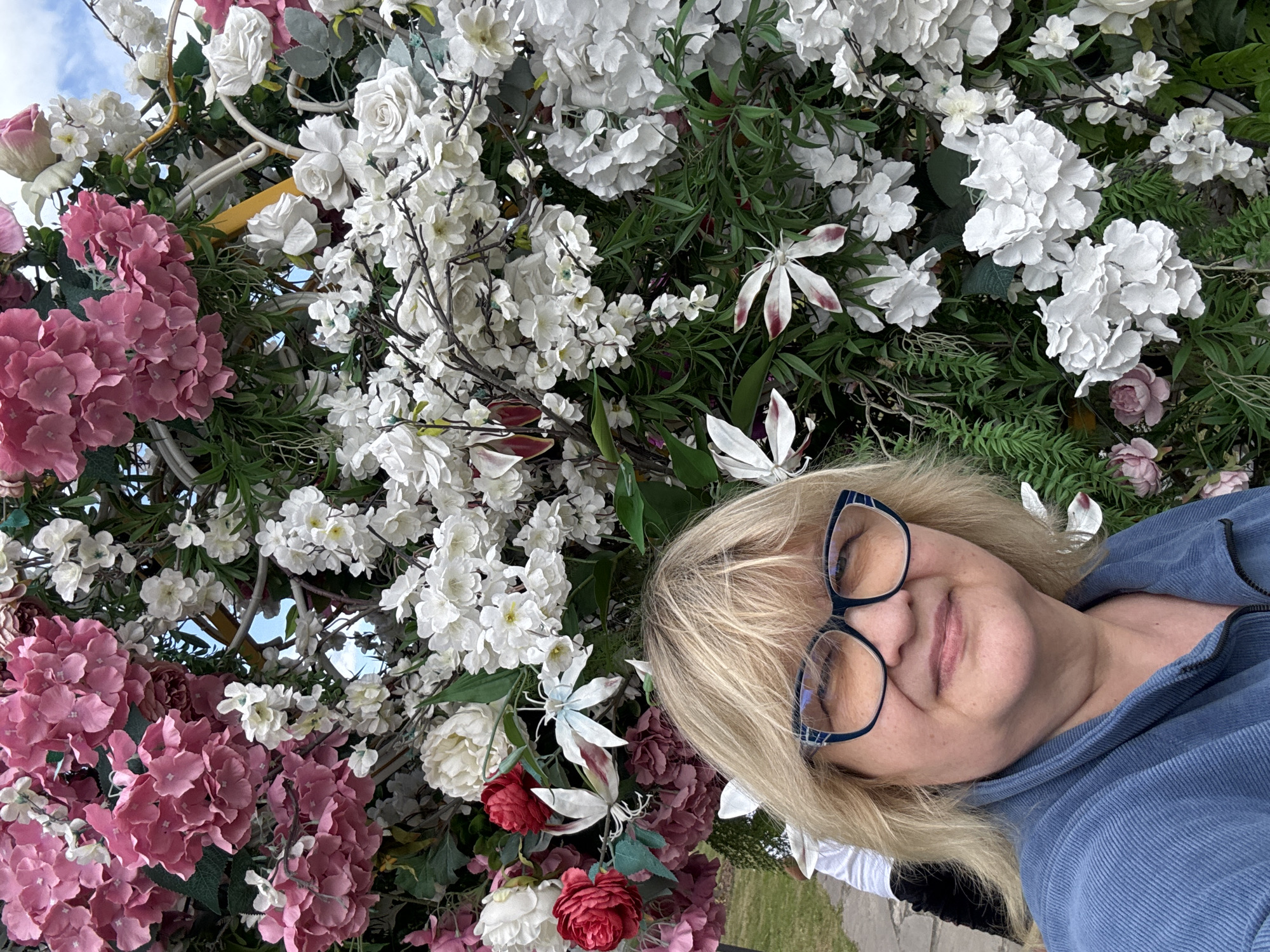 A medium-shot, eye-level, outdoor, natural light, selfie captures a fair-skinned woman with blonde hair, wearing large blue-rimmed glasses and a blue corduroy jacket. She is smiling and positioned in front of a large, elaborate floral arrangement. The arrangement is densely packed with artificial flowers, primarily in shades of pink and white, with some red and darker pink accents. Green foliage is interspersed throughout the flowers, creating a lush backdrop. The woman's head and shoulders are the primary focus, with the floral display filling the rest of the frame. The background behind the woman and the flowers shows hints of a paved walkway and possibly some green grass and a cloudy sky, suggesting an outdoor setting. The specific location in the city of Serakseyevo, Russia, cannot be determined from the visual information alone.