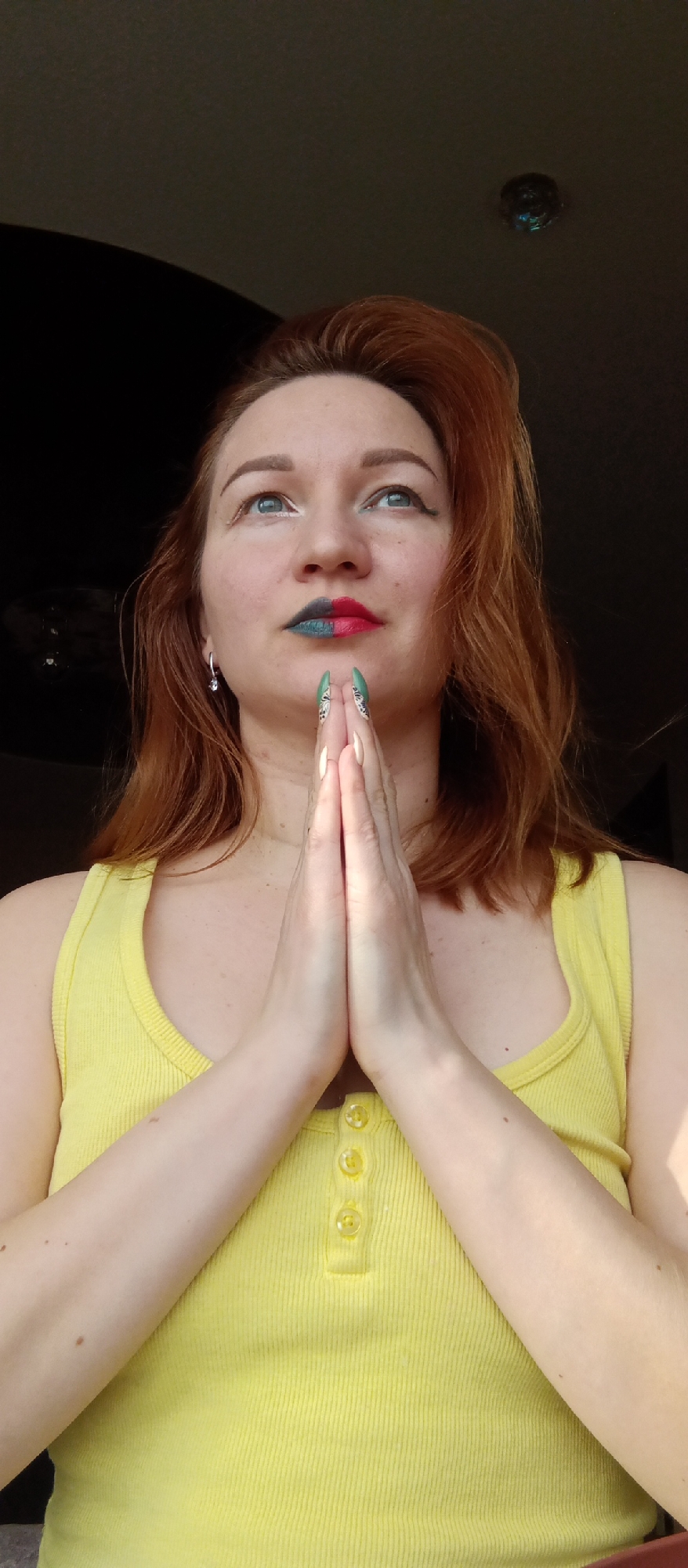 A woman with red hair, wearing a yellow tank top, is seen from a low-angle perspective indoors in Unknown, Unknown. Her hands are pressed together in front of her in a prayer-like gesture. She has a two-toned lipstick, with one side of her lips painted dark blue and the other side painted red. She is looking upwards. There is a light fixture on the ceiling.