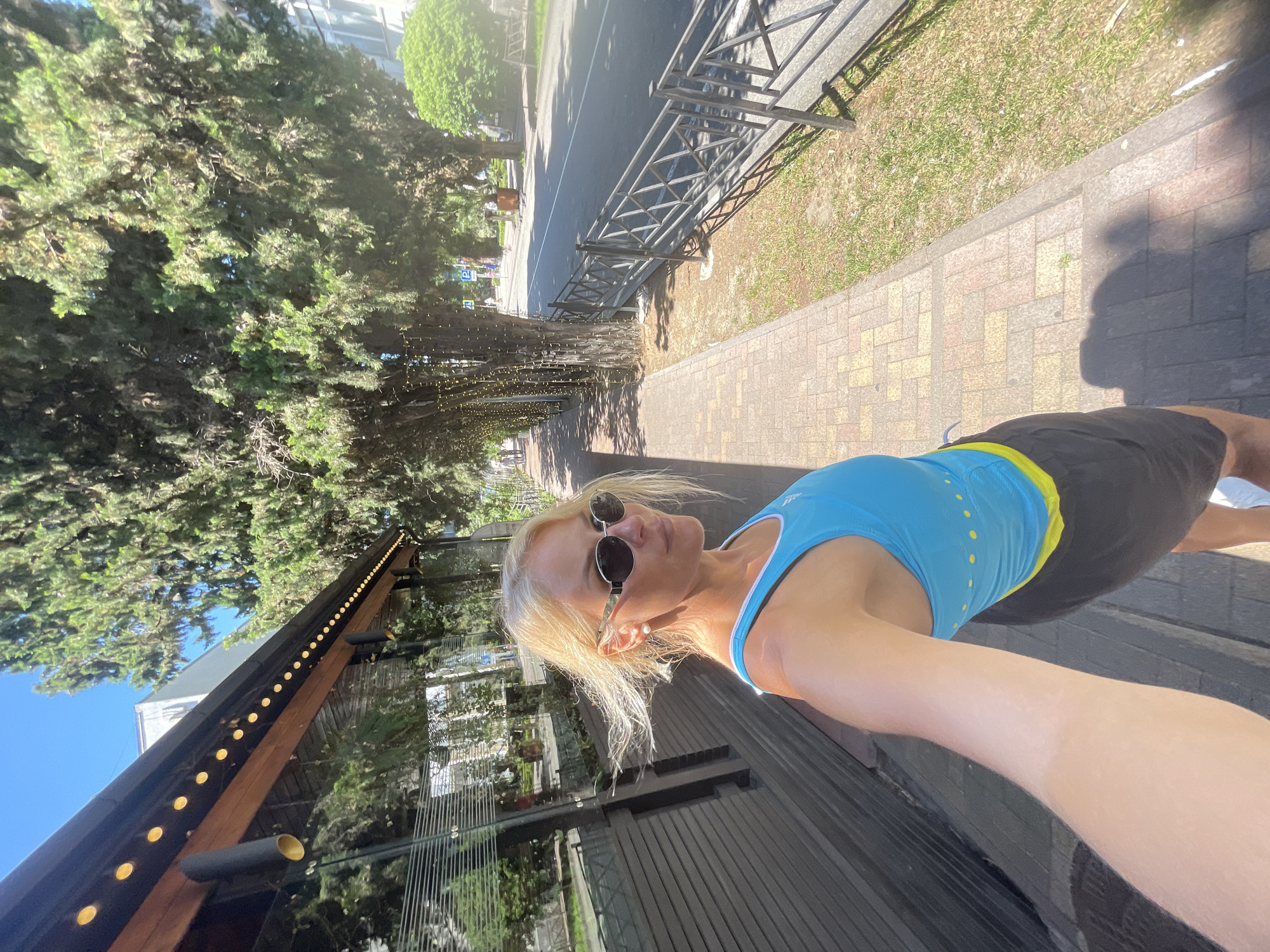 A blonde woman with sunglasses and a blue tank top is taking a selfie on a sunny day in Sochi, Russia. She is standing on a brick sidewalk next to a building with large windows and a row of tall trees along the street. The trees are decorated with string lights. A black metal fence runs along the side of the road, and a car is visible driving on the street. The woman is wearing black shorts.