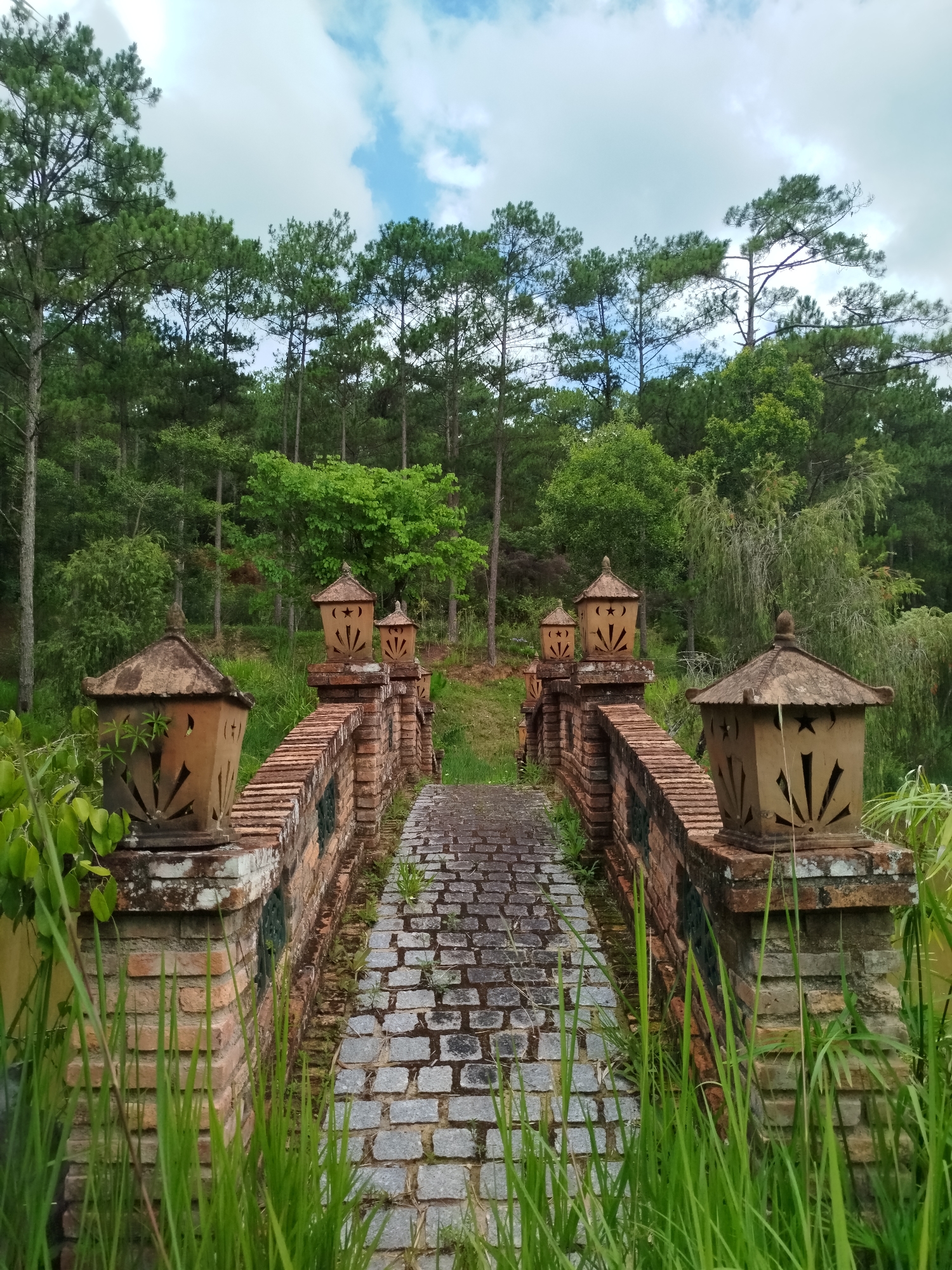 A long, slightly elevated bridge constructed of grey cobblestone spans a grassy terrain towards a dense forest. The bridge features low brick walls on either side, punctuated by ornamental, lantern-like structures on brick pedestals. The lanterns are a light-brown hue with star and sun-shaped cutouts. Green grass grows abundantly around the base of the bridge. The forest in the background is composed of a mix of deciduous trees and tall, slender conifers under a sky with puffy clouds. The location is Cam Ly - Da Lat, Vietnam.