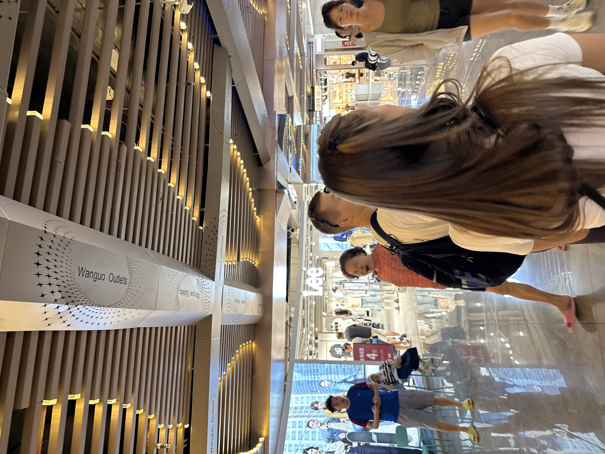 This image captures a scene inside a shopping mall, likely in the Haizhu District of China, indicated by the "Wangguo Outlets" signage visible on the ceiling structure. In the foreground, a woman with long, dark brown hair and a white t-shirt is viewed from behind, facing away from the camera. To her left, a man with short brown hair, wearing a blue t-shirt and grey shorts, stands looking towards the camera. Further into the mall, a woman in an orange-patterned top is captured mid-yawn, facing right. Behind her, a man in a black t-shirt and shorts walks away from the camera. Several clothing racks and mannequins are visible in the background, with the "Lee" logo prominently displayed on a storefront. The mall features a modern ceiling design with horizontal wooden slats and recessed lighting, and a polished, reflective floor. Large digital screens display images of people. The overall atmosphere appears to be a busy commercial environment.