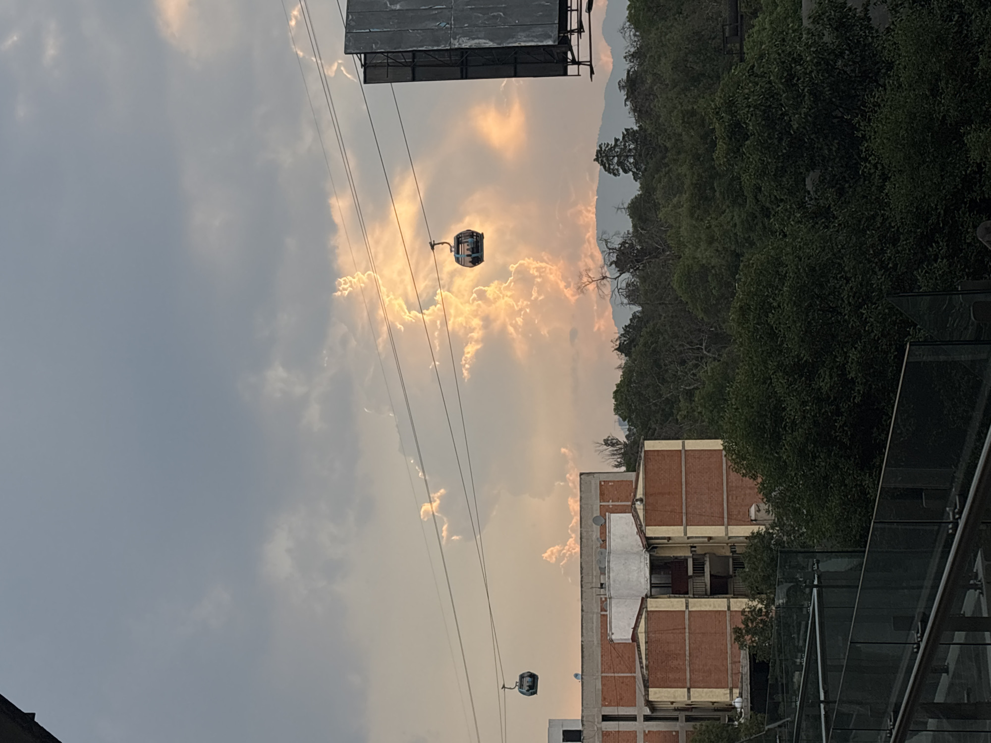 The image depicts a daytime scene in Mexico City, Mexico, featuring a cable car system. Two cable cars are visible, suspended by wires against a backdrop of cloudy sky and buildings. The sky is partially illuminated with a golden hue, suggesting sunset or sunrise. Dense green foliage and trees are visible on the right side of the image, contrasting with the urban elements. The architecture includes brick buildings and a glass structure in the foreground.