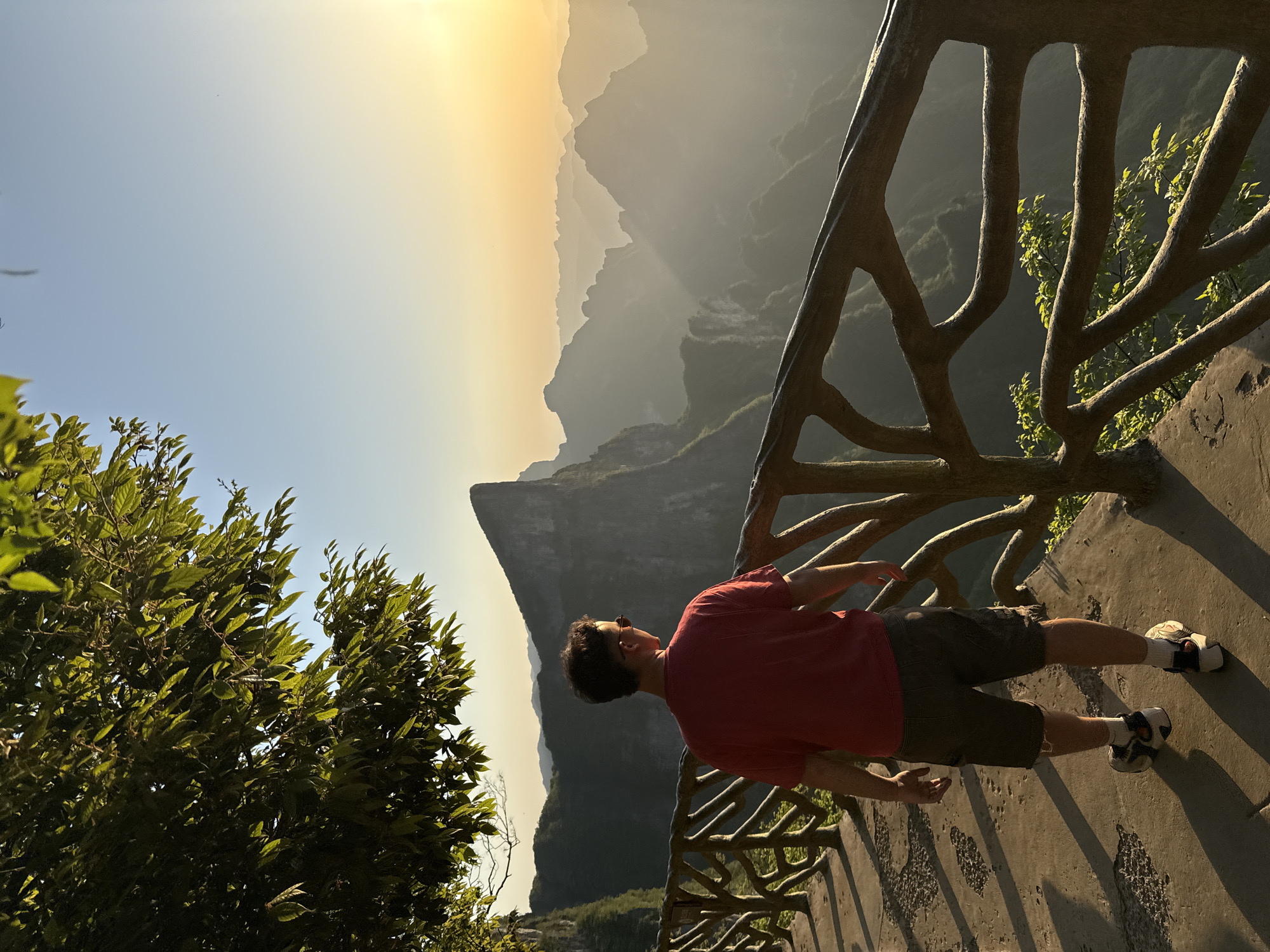 A man stands on a stone observation deck, leaning against a wooden railing overlooking the mountains. He wears a red short-sleeved shirt and dark shorts, sneakers, and dark sunglasses. His gaze is directed towards the mountains. The view includes a steep cliff, with many mountains in the distance shrouded in haze, illuminated by the setting sun. The photo was taken in Yongding District, China. A tree grows in the foreground.