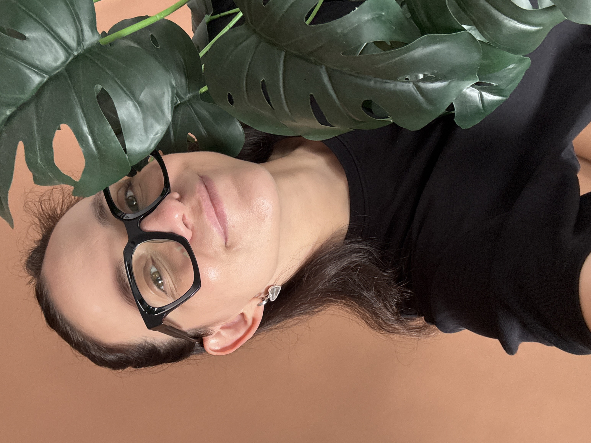 A close-up, upward-angle shot shows a woman with dark hair and eyeglasses smiling. She is wearing a black t-shirt and a silver heart-shaped earring. A large, green monstera plant is partially obscuring her face. The background is a solid, light brown color. The image does not contain information about the location in Chaikovsky, Russia.