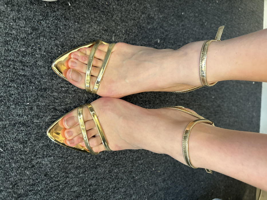 A close-up, top-down view captures a pair of human feet, presumably female, standing side-by-side on a dark gray, short-pile carpet. The scene is located indoors in Arcueil, France. Both feet are adorned with golden, pointed-toe, open-toed high-heeled sandals. Each sandal features a thin strap secured around the ankle and two parallel straps across the top of the forefoot, exposing the toes. The skin tone of the feet is fair. The toenails are visible and appear natural, unpolished, or with a clear coat; the great toenail on the left foot (viewer's left) exhibits a slightly thicker or discolored appearance. The carpet shows a consistent texture across the visible area. A light-colored, possibly white, wall or baseboard is faintly visible at the very top edge of the frame.