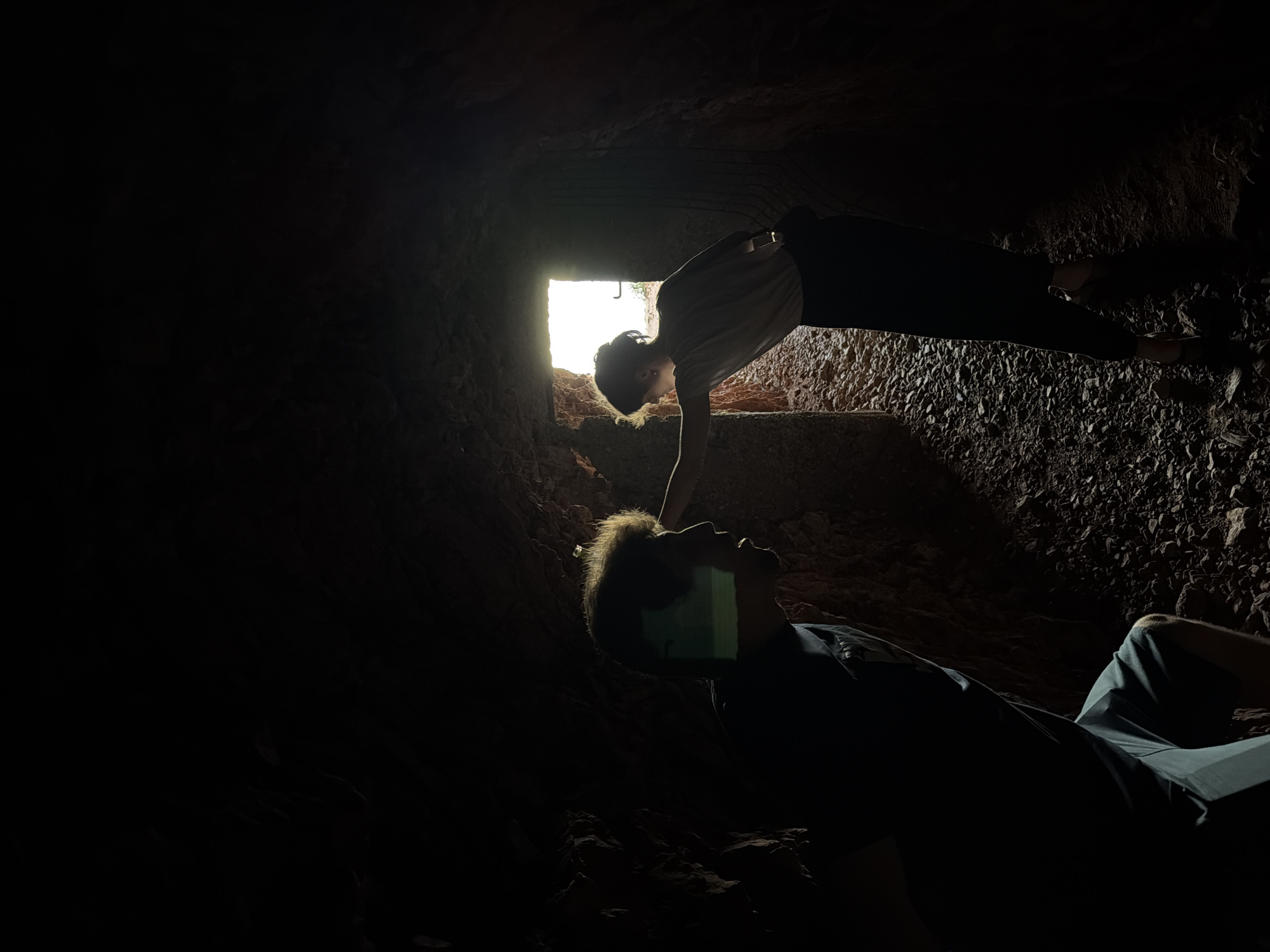 The image depicts two individuals inside a dimly lit, rough-hewn tunnel or cave. The tunnel walls appear to be made of reddish-brown earth and rock, with a rough, uneven texture. The ground is covered in loose rocks and gravel.
In the foreground, on the left side of the image, is a person silhouetted against the darkness, facing to the right. Their head is turned and their mouth is slightly open. They are wearing a dark t-shirt and light-colored shorts. A bright green band is visible on the back of their head.
Further into the tunnel, towards a bright opening, stands a second person. This individual is also silhouetted, leaning towards the opening and appearing to be looking out. They are wearing a light-colored t-shirt and dark pants. Their hair is pulled up, and they have earrings.
The opening at the end of the tunnel emits a strong, bright light, suggesting an exit to the outside. The contrast between the dark interior and the bright exterior is significant. The overall impression is one of exploration or passage through a natural or man-made subterranean space. The location is identified as Petrovac centar, Montenegro.