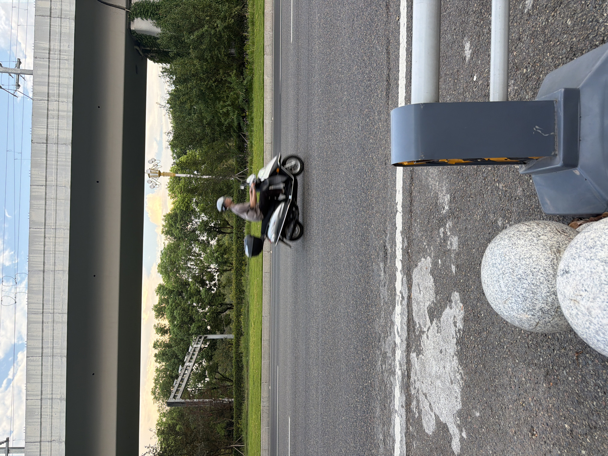 The image shows a road in Jiangjin, China, with an elevated highway above. A person on a scooter is riding on the road, which is lined with greenery and trees. In the foreground, there are decorative spherical stones and a gray barrier. The sky is visible above the elevated highway, with some clouds. The scene appears to be set during the day, possibly in the late afternoon or early evening, given the lighting conditions.