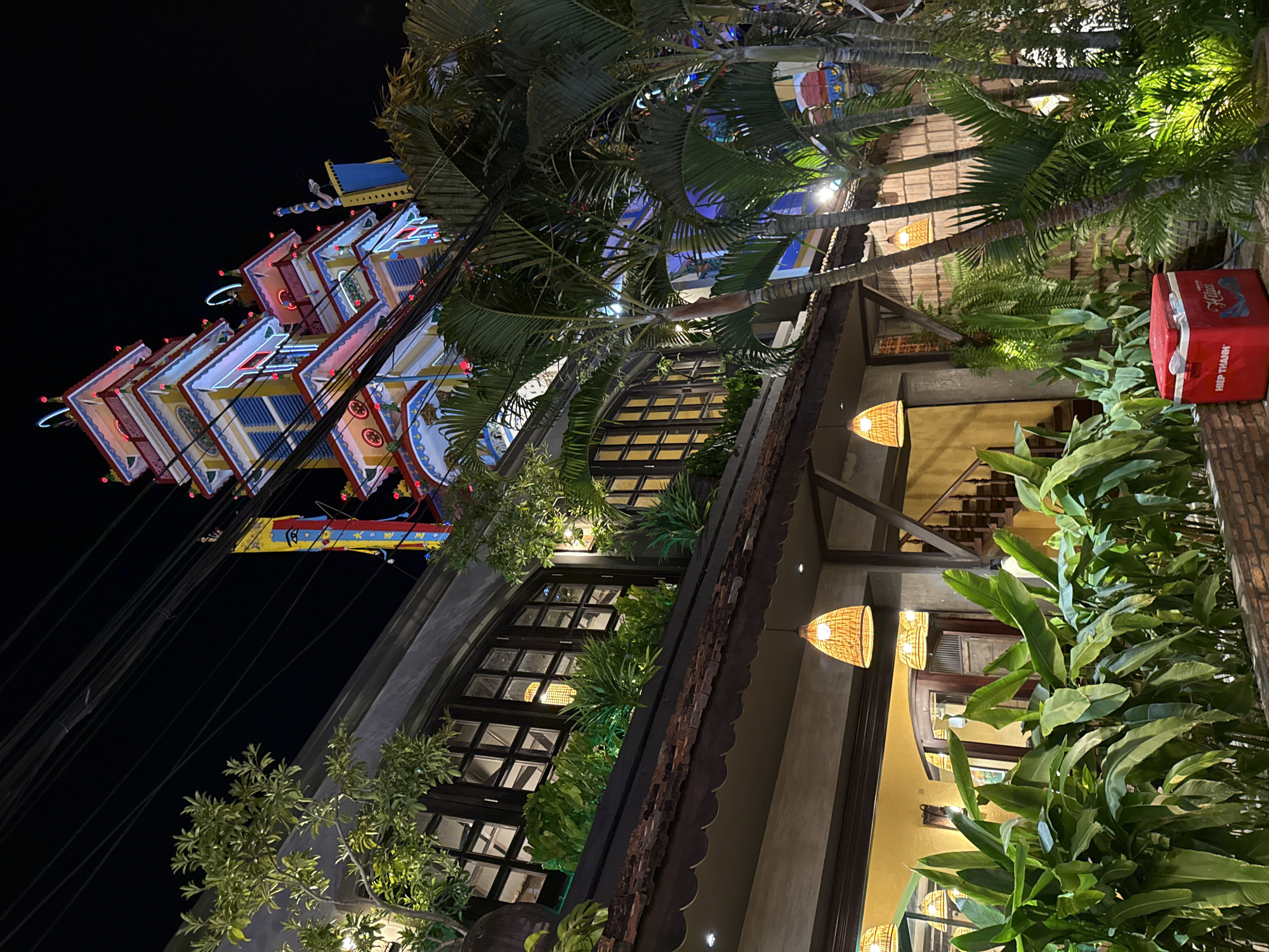 This image depicts a night scene outside a building in Nha Trang, Vietnam. The building features a multi-tiered, brightly lit tower structure in the background, adorned with colorful decorations and what appear to be neon lights. In the foreground, there is a covered outdoor area with large windows, plants, and woven hanging lamps. Palm trees and other lush greenery are visible, illuminated by artificial lighting. A red cooler box with text that reads "HIỆP THANH HILUX" is placed on the ground in the lower right corner. The environment appears to be an outdoor dining or seating area, possibly a restaurant or cafe, given the ambiance and the presence of the cooler. The sky is dark, indicating it is nighttime.