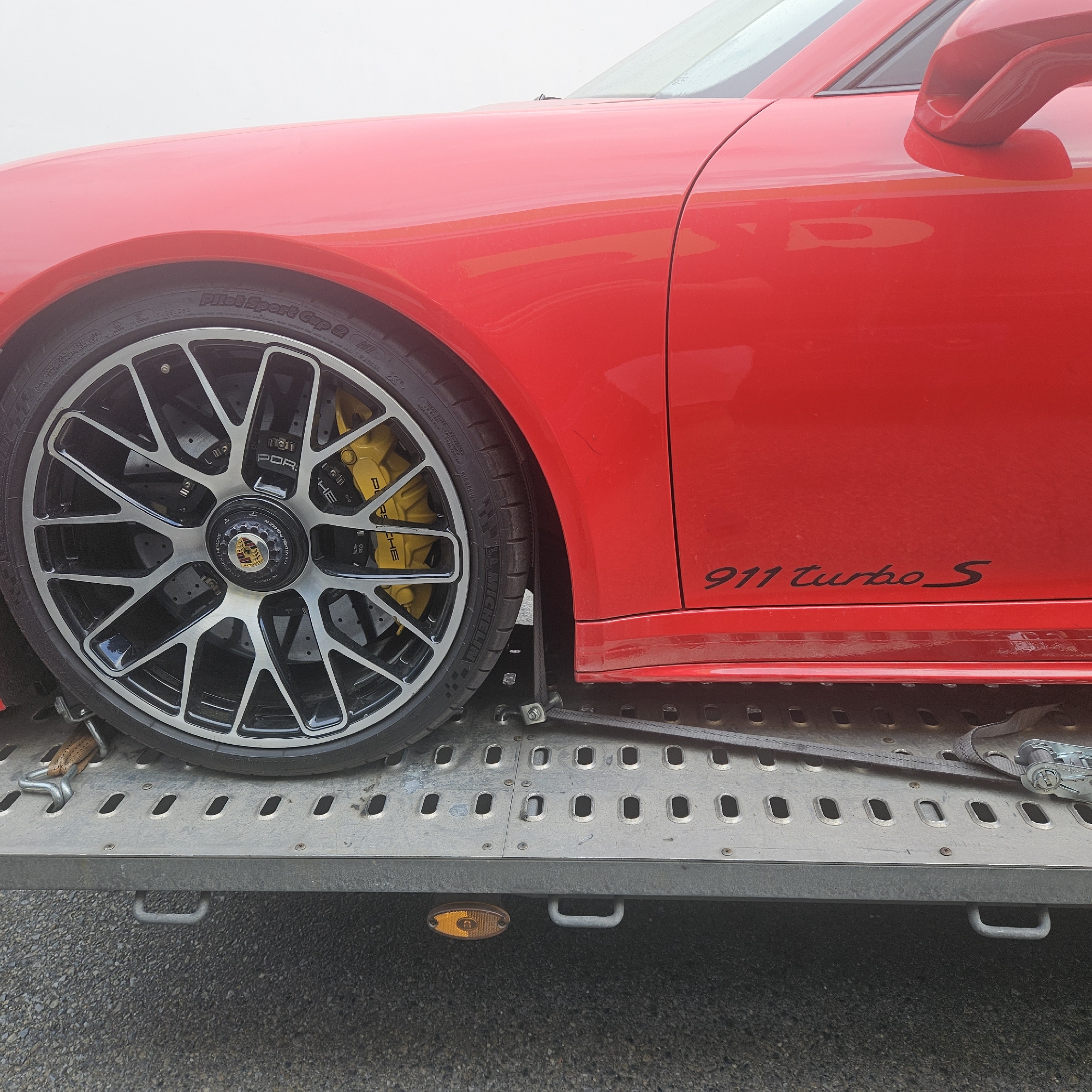 The image shows a close-up view of the front passenger side wheel and fender of a red Porsche 911 Turbo S. The car is parked on a metal car carrier trailer, secured with straps. The wheel is a multi-spoke alloy design with yellow brake calipers visible behind it. The tire has "Michelin Pilot Sport Cup 2" branding. The side of the car features the "911 Turbo S" badge. The background is a plain white surface, likely the sky or a studio backdrop. The scene is static and depicts a vehicle being transported. The location is identified as La Jonquera, Spain.