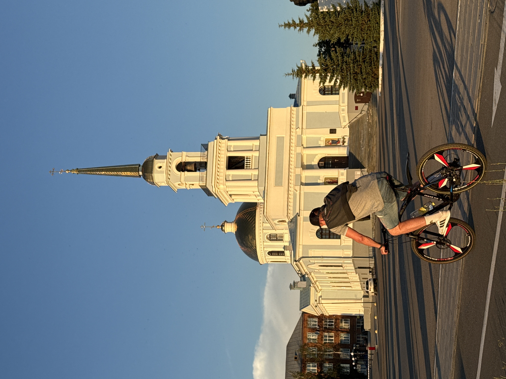 This is a full-frame, eye-level, outdoor, vertical shot of a person cycling on a paved road in front of a large, light-blue and white church with a golden dome and spire. The sun is setting, casting long shadows. The church appears to be the Cathedral of the Assumption of the Blessed Virgin Mary, located in Votkinsk, Russia. The cyclist, a man wearing a dark baseball cap, grey t-shirt, and denim shorts, is moving away from the camera. The bicycle has black wheels with red and white accents. In the background, to the left of the church, is a multi-story brick building. Two evergreen trees stand to the right of the church's entrance. The sky is clear and blue.