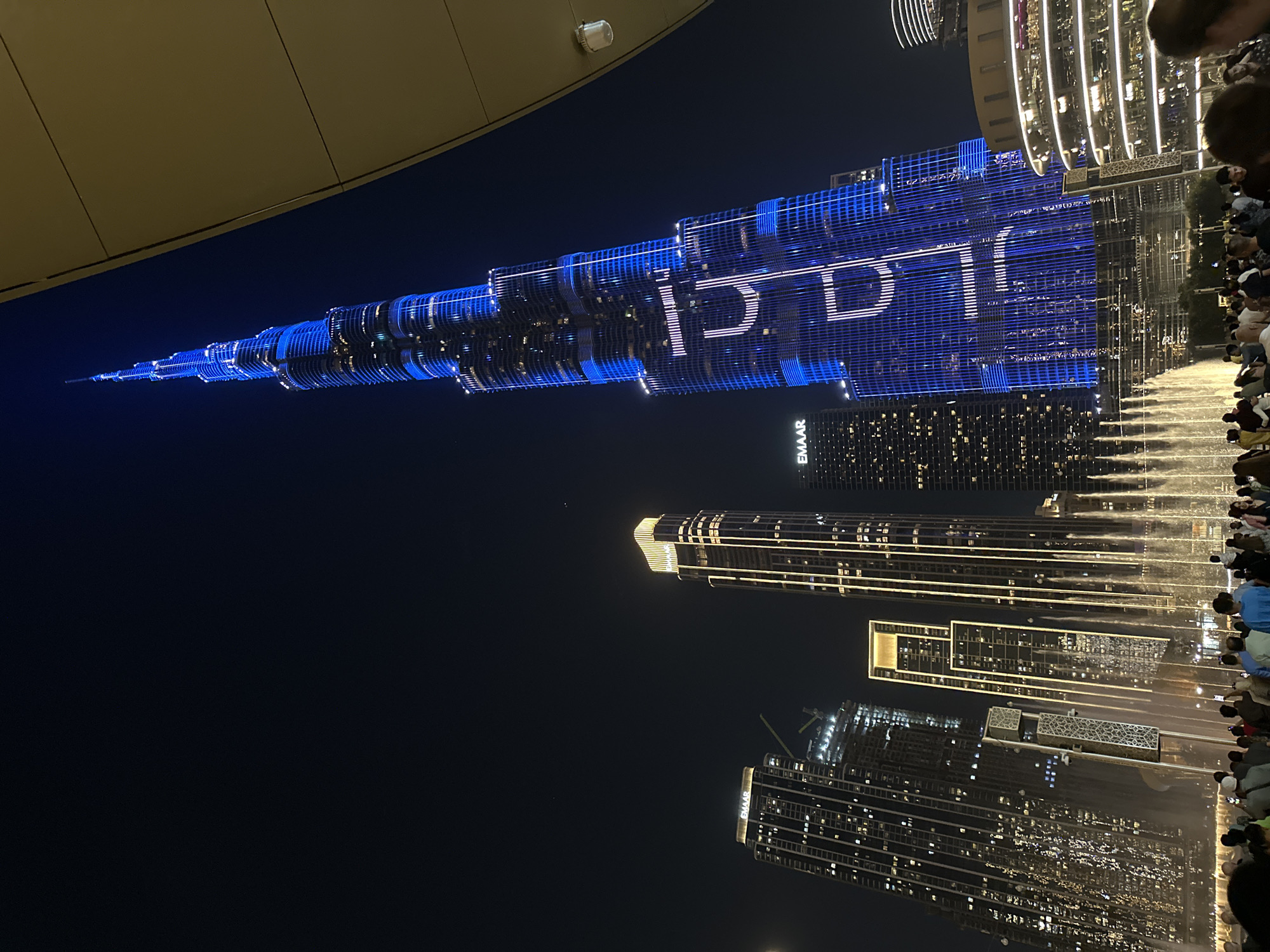 The image presents a spectacular night view of the Burj Khalifa in Dubai, United Arab Emirates, illuminated by a dynamic light and projection show. The towering skyscraper is aglow with vibrant blue lights and intricate white patterns, prominently featuring large white Arabic text that appears to spell "دبي" (Dubai) vertically along its facade. Below this, other white lights create additional design elements.
In the mid-ground, the famous Dubai Fountain is active, with bright, illuminated water jets rising from the basin. A crowd of people is gathered along the edge of the fountain, their silhouettes visible as they gaze upon the synchronized light and water performance.
Surrounding the main attractions are numerous other modern high-rise buildings, their windows and exteriors illuminated against the dark night sky. The "EMAAR" logo is clearly visible on one of the buildings, indicating the developer. Part of a large, beige-colored building or structure is visible in the upper left foreground. The clear night sky enhances the brightness of the urban lights. The scene captures the lively atmosphere of a major tourist attraction, with spectators enjoying the impressive architectural and artistic display.