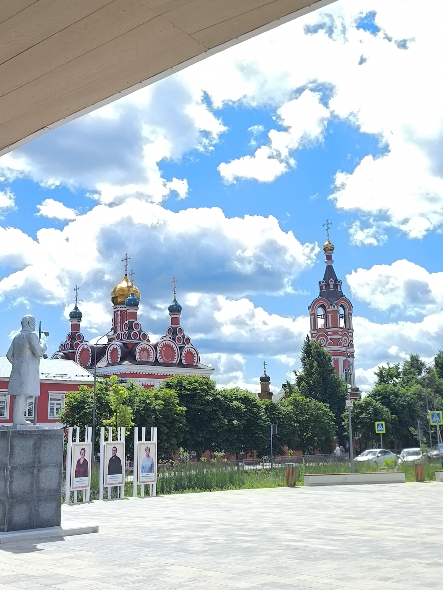 The image captures a view of the Church of the Transfiguration of the Lord in Taldom, Russia, on a partly cloudy day. In the foreground, a stone statue of a man holding a cross stands to the left, on a stone pedestal. Behind the statue, a line of three portraits on white stands displays individuals with their names. The church, with its red and white facade and golden domes, is visible in the middle ground, flanked by green trees. To the right of the church is a red and white bell tower topped with a golden dome and a cross. The background shows a street with cars and traffic signs, suggesting an urban setting. The sky is a vibrant blue with scattered white clouds.