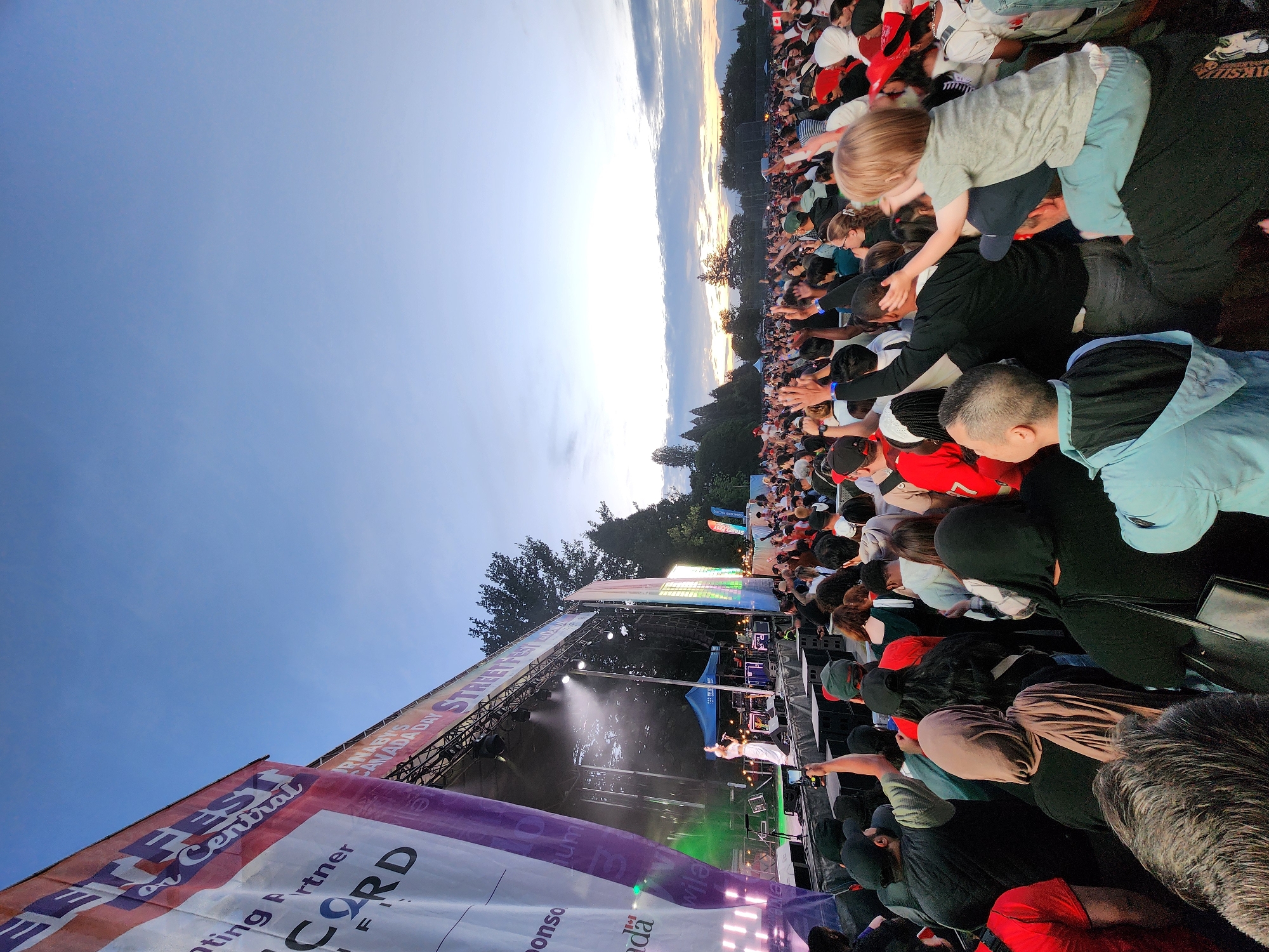 A wide-angle, low-angle outdoor photograph captures a lively concert scene with a large crowd of people gathered in front of a brightly lit stage. The sky above is a gradient of blue and orange, indicating either dawn or dusk, with soft clouds. Dominating the left side of the frame is a large banner with text that includes "STREETFEST" and "CANADA DAY." On stage, a female performer in a white outfit is singing into a microphone, illuminated by spotlights. The crowd is diverse, with many individuals raising their hands and appearing to be enjoying the performance. Some people are visible from the chest up, while others are shown from the waist up, and in the foreground, some individuals are seen from the back. The environment suggests a public park or open venue, with trees visible in the background. The overall atmosphere is festive and energetic.