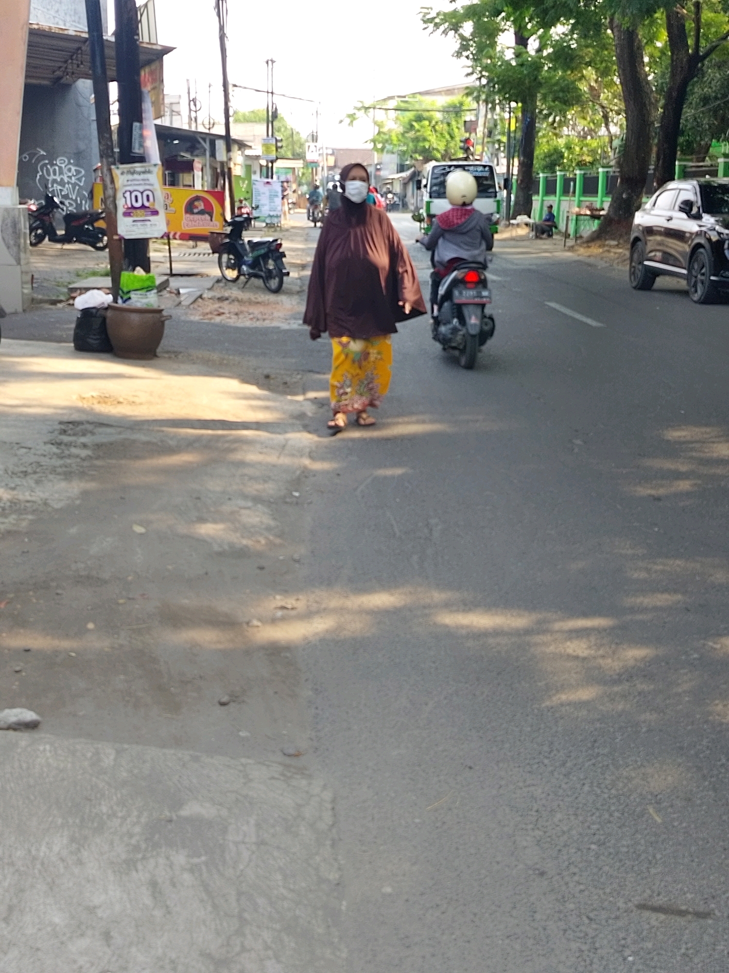 The image captures a street scene in Mojokerto, Indonesia, likely during daytime. In the foreground, a woman wearing a brown hijab and a yellow patterned skirt walks on a paved road. She is also wearing a white face mask. To her left, a dark-colored car is parked on the side of the road. Further down the road, another person is riding a scooter. The background shows buildings, trees, and more vehicles, suggesting a typical urban environment. The overall impression is a snapshot of everyday life on a street in Mojokerto.