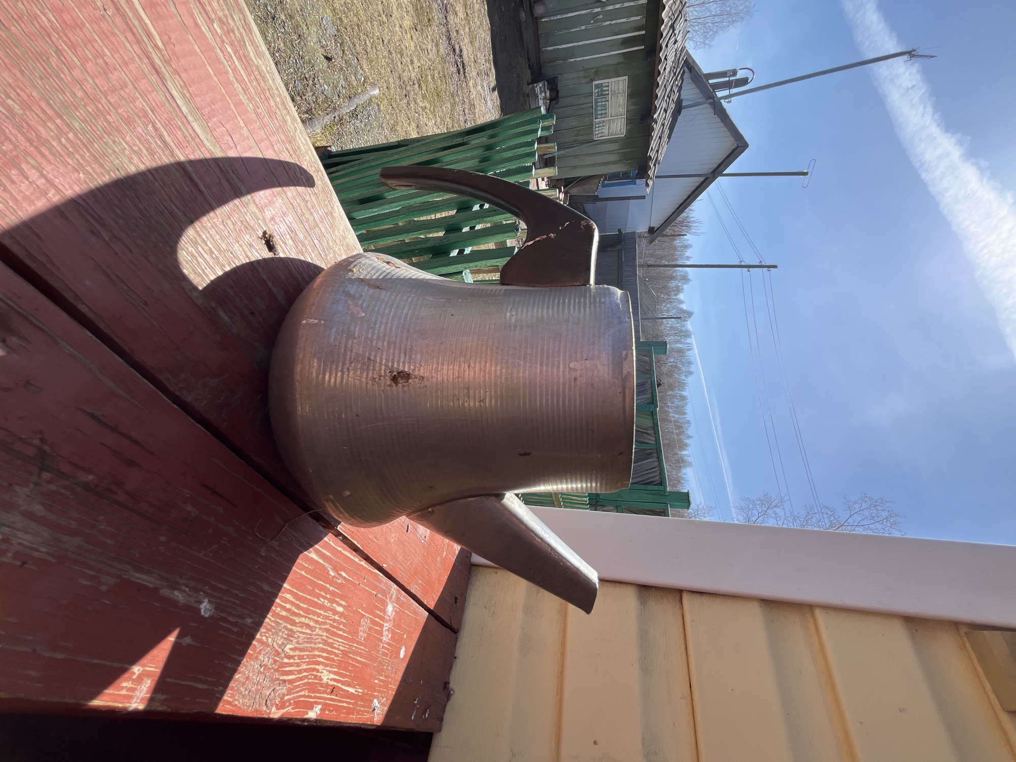 A tarnished metal pitcher with a handle and spout sits on a weathered wooden surface painted red in an outdoor setting. The scene includes a green picket fence, a building with a gray roof, and clear blue sky with a contrail. The location is the city of Верхняя Уря, Russia.