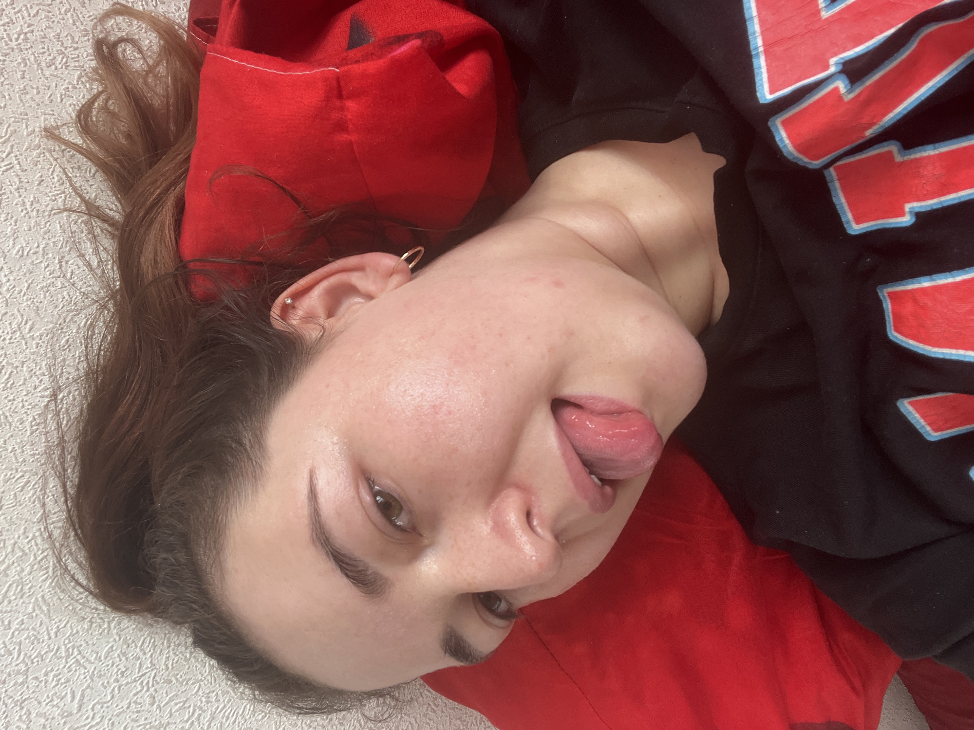 The image shows a close-up, high-angle, selfie-style shot of a young woman lying down. She has brown hair and is wearing a black t-shirt with red and blue lettering. Her tongue is sticking out. She is lying on a red pillow against a textured white wall. There is no indication of the city of Chelyabinsk, Russia, or any other specific location in this image.