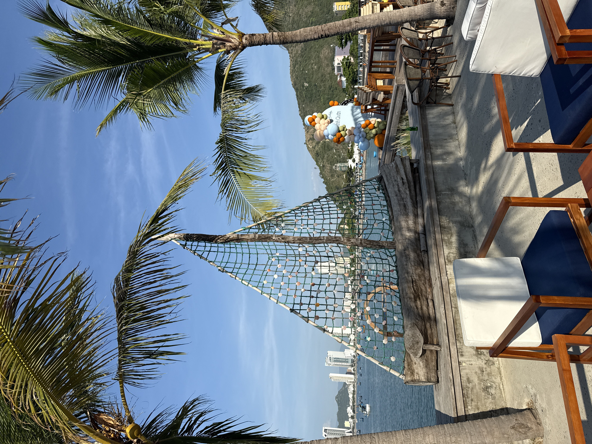 The image captures a sunny outdoor seating area near the coast of Bac Nha Trang Ward, Vietnam. In the foreground, two wooden chairs with dark blue cushions and white backrests are arranged on a concrete patio. To the left, a portion of a palm tree trunk is visible. In the background, a decorative sail made of netting and shells is positioned on a wooden platform overlooking the sea. The sea itself is a calm blue, with several boats scattered across its surface. Beyond the water, a city skyline with several modern buildings and a distant mountain range can be seen. To the right of the sail, a cluster of colorful balloons forms a festive decoration, with a circular sign bearing text that appears to be Vietnamese. Further in the background, more seating areas with wooden tables and chairs are visible, some under the shade of palm trees. The sky is a clear, bright blue with a few wispy clouds. The overall scene suggests a relaxed, tropical atmosphere, likely a resort or restaurant setting. There are no people visible in the image.