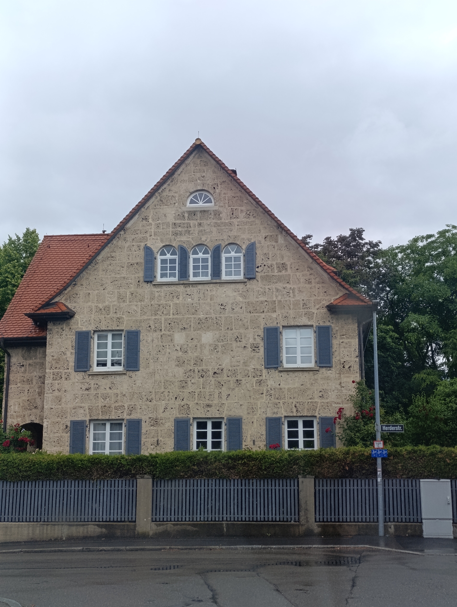 The image shows a two-story stone house with a red tile roof, typical of European architecture, situated in Reutlingen, Germany. The house has several windows with blue shutters, a small arched window on the upper gable, and a red-tiled mansard roof. A dense green hedge runs along the front of the property, behind a gray slatted fence. A street sign indicating "Herdersstr." is visible on a lamppost, confirming the location. The sky is overcast, suggesting a cloudy day, and the street in front of the house appears wet, possibly from recent rain. The overall scene depicts a residential street in a European city.