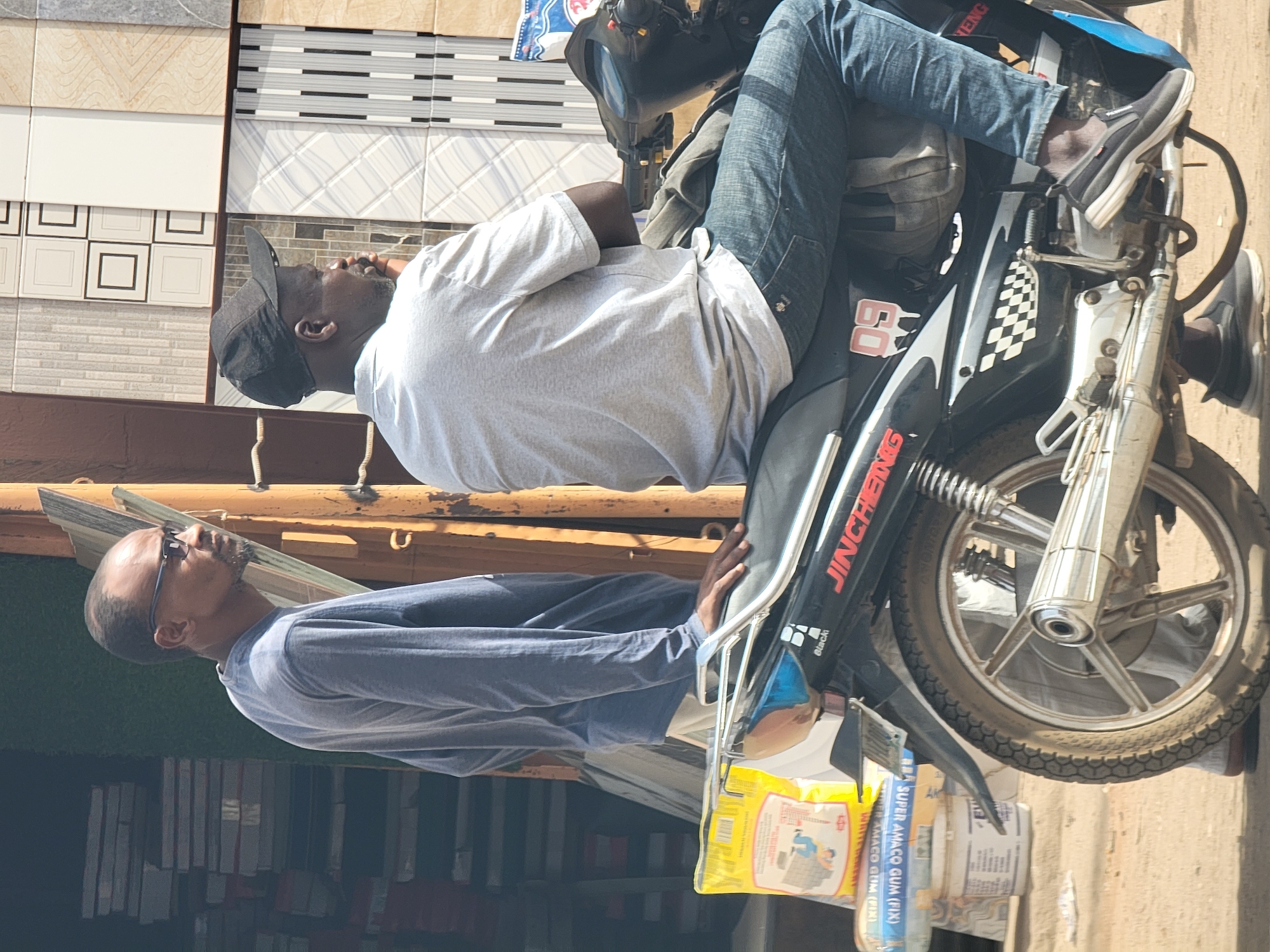 A man wearing a grey t-shirt, blue jeans, and black sneakers straddles a black motorcycle with the word "JINCHIEING" printed on its side. A second man wearing glasses and a long-sleeved blue shirt stands nearby. The motorcycle has a front rack with packages of what appears to be "SUPER AMACO GUM (FIX)". In the background, there is a display of tiles, indicating the setting could be a business related to construction or home improvement. The image appears to be taken in Maiburiji, Nigeria, based on the product packaging.