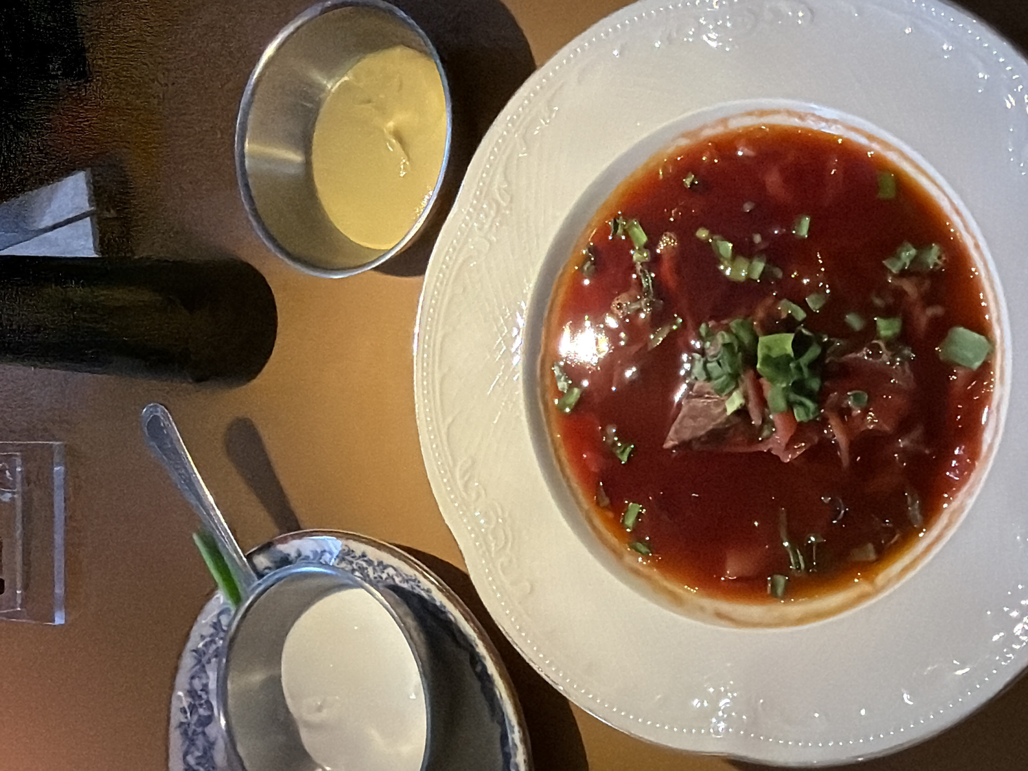 The image presents an overhead view of a dining setting, likely in a restaurant or cafe in Krasnoyarsk, Russia. The central focus is a large, white ceramic bowl filled with a rich, deep red soup, characteristic of borscht. The soup is generously garnished with chopped green herbs, resembling green onions or dill, and contains visible pieces of meat or beet. The white bowl features an elegant, embossed pattern along its wide rim.
Adjacent to the soup bowl, in the upper left, sits a small, shiny metallic cup containing a pale yellow, creamy condiment, potentially sour cream or mustard, which are traditional accompaniments for borscht. Below the main soup bowl, a smaller, decorative cup or bowl with a blue and white intricate pattern is partially visible, holding a silver spoon. On the far left, a dark, cylindrical object, possibly a salt or pepper shaker or napkin holder, and a clear, rectangular item are present on the brown, textured tabletop.
The scene depicts a meal being served or awaiting consumption. No people are visible. The lighting is warm and appears to be indoor, not indicating a specific time of day. There is no visible text in the image. The presentation of the dish and accompaniments suggests a typical meal in a Russian establishment.