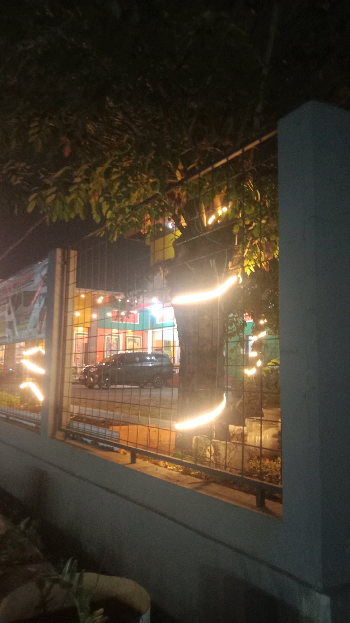 The image depicts a nighttime scene behind a metal fence in Satriajaya, Indonesia. A dark-colored SUV is parked in front of a building with colorful walls, likely a shop or restaurant, illuminated by warm-toned lights. String lights are visible, draped around trees and along the fence, creating a festive atmosphere. The foreground shows a concrete wall and some greenery. The overall impression is of a vibrant, possibly commercial area at night.