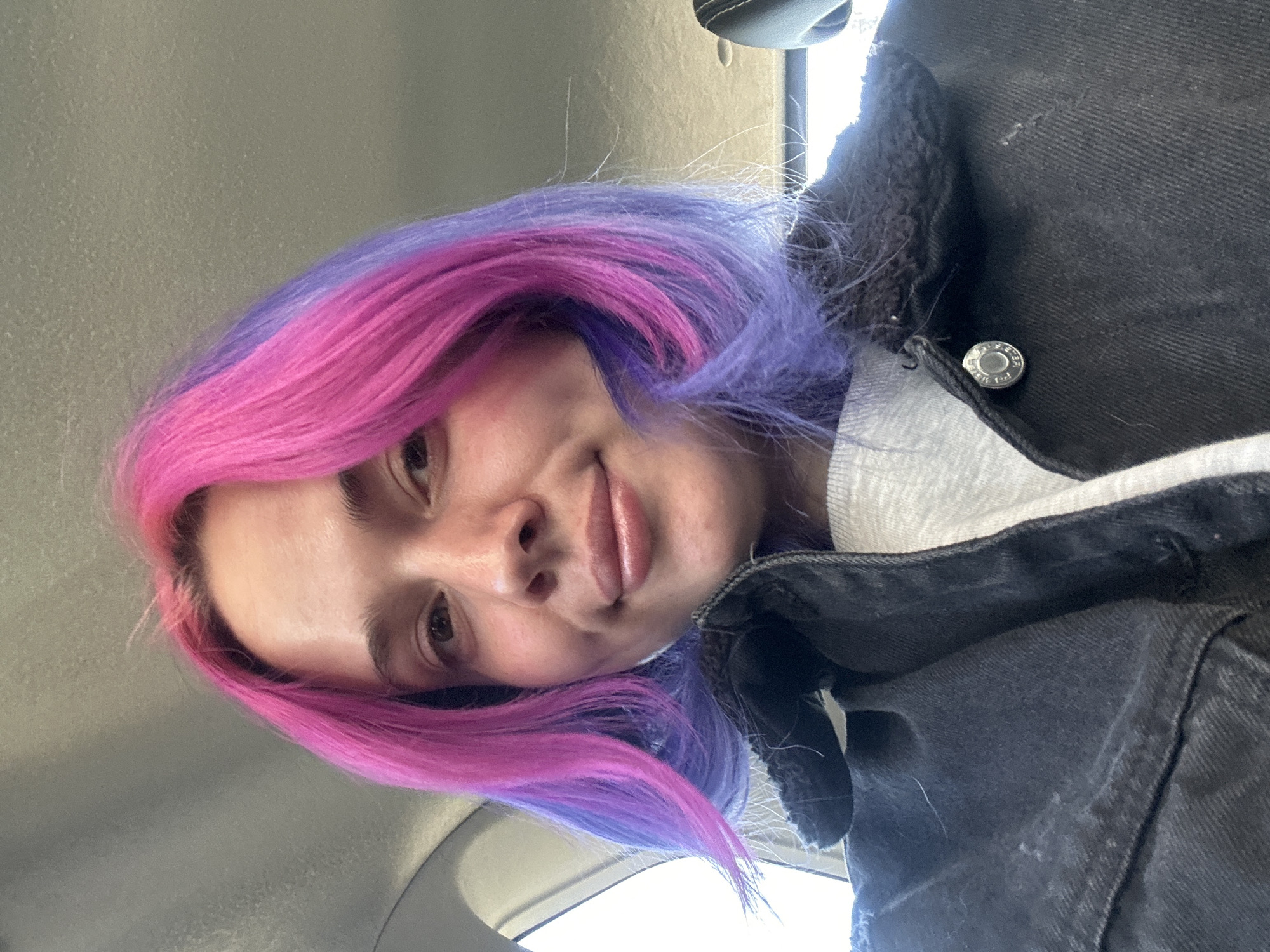 The image displays a close-up, low-angle portrait of a young person. They have strikingly vibrant, two-toned hair, with fuchsia pink on one side and rich purple on the other, styled in a medium-length cut. The person is looking directly at the camera with a gentle, soft smile, suggesting a pleasant and calm mood.
They are wearing a dark denim jacket with a visible textured, possibly sherpa, lining around the collar and edges. A light-colored hoodie or top is layered underneath. A metallic button on the jacket is visible, though any text embossed on it is too faint and small to be legible.
The setting appears to be indoors, specifically within a vehicle, indicated by the light-colored interior ceiling and blurred elements in the background, consistent with a car's cabin. The lighting is bright and natural, suggesting the photo was taken during the daytime. The overall impression is a casual, relaxed selfie or portrait.