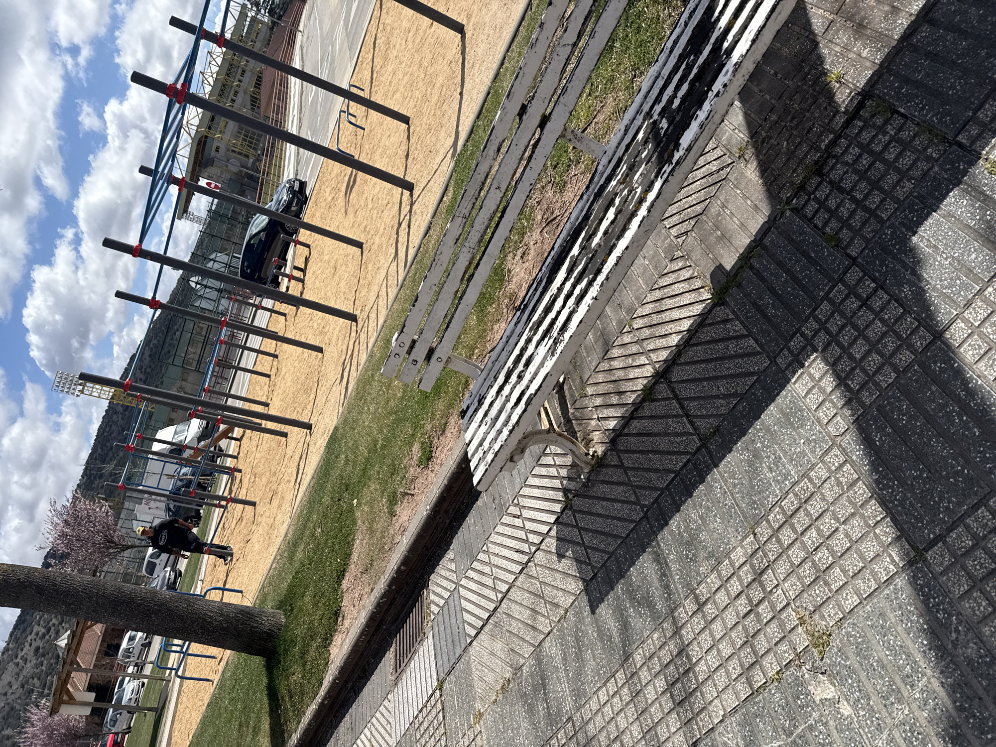 The image depicts an outdoor fitness area in Soria, Spain, with various exercise structures, including horizontal bars. The ground is a mix of sand and grass. A white wooden bench sits adjacent to a tiled walkway. A tree trunk is visible to the left of the frame. In the background, there are buildings and a hillside. The sky is partly cloudy. People are visible using the fitness equipment.
