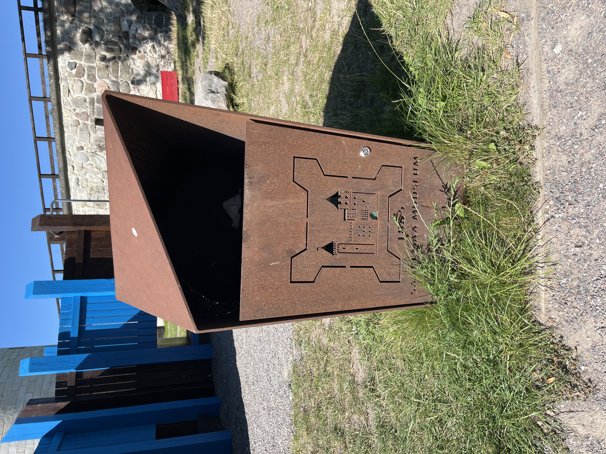 A rusted metal trash receptacle with a crest of a castle and the text "SAAREMAA MUSEUM" is positioned on a grassy area with gravel and sand under a clear blue sky. In the background, a stone wall with a wooden railing runs along the top, and a section of blue and brown wooden fencing is visible to the left. The trash receptacle has an angled opening at the top, and a lock mechanism is present on its side. The scene appears to be outdoors, possibly in a park or historical site.