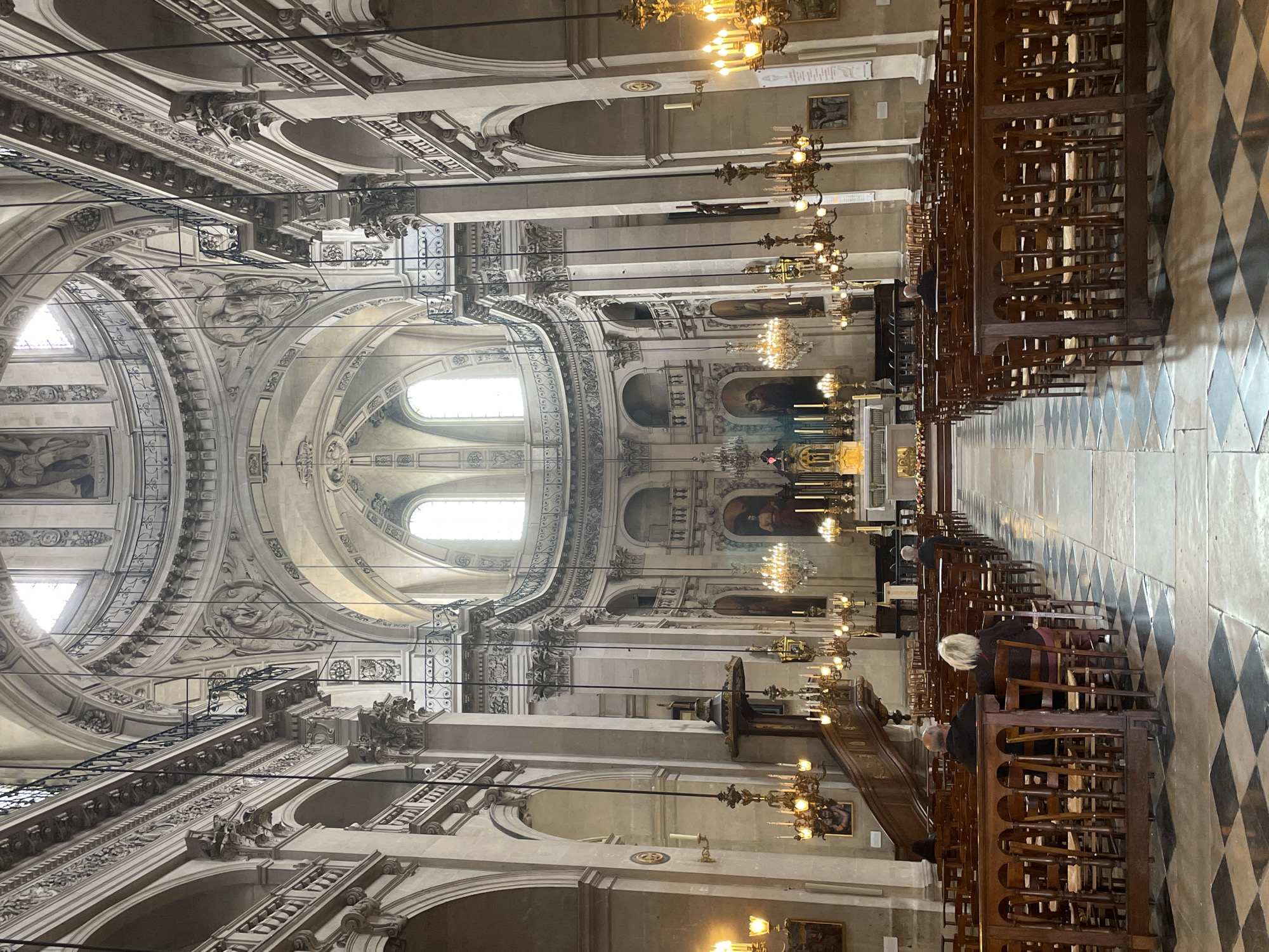 The image depicts the interior of a grand cathedral in Paris, France, likely Saint-Paul-Saint-Louis Church, given its architectural style and dome. The perspective is from the rear of the nave, looking towards the altar. Rows of empty wooden pews with dark wooden dividers fill the foreground and extend down the length of the nave. Several individuals are seated in the pews, facing towards the front.
The cathedral's architecture is ornate, with high vaulted ceilings, intricate carvings, large arched windows allowing natural light to filter in, and classical columns. Several large, multi-branched chandeliers with flickering candle-like bulbs hang from the ceiling, providing warm, ambient lighting. Paintings adorn the walls, and a prominent wooden pulpit is visible on the left side. The floor is paved with a checkerboard pattern of black and light-colored tiles. The overall atmosphere is one of solemnity and reverence.