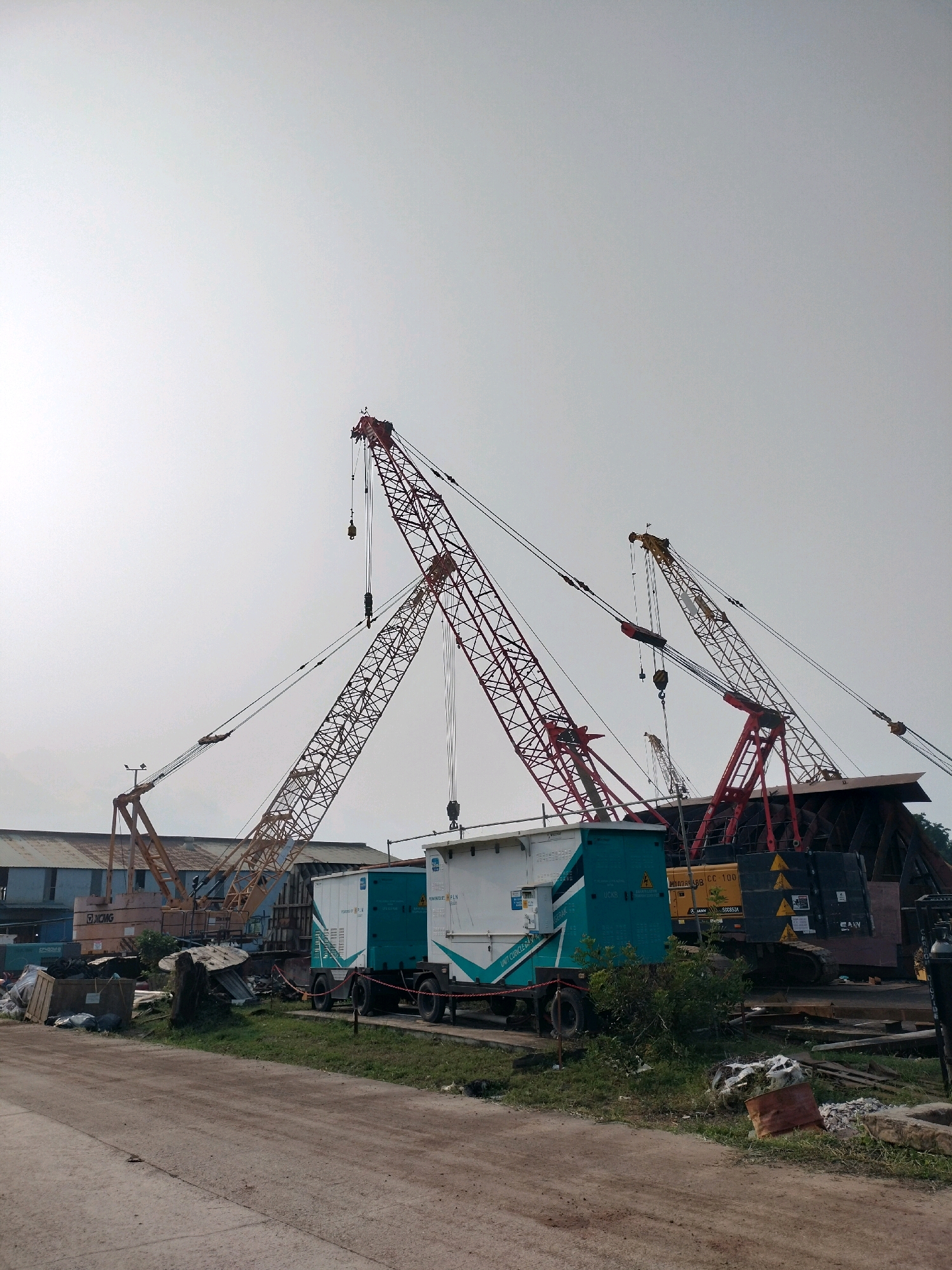 This image shows a construction site with multiple cranes and portable generators in Batam, Indonesia. The foreground features a dirt road with grass and debris. In the mid-ground, there are two portable generators, one teal and white and the other white with teal accents, situated on trailers. Behind these, several large construction cranes dominate the scene, their red and yellow lattice structures reaching high into the sky. A large, dark, metal structure, possibly part of a ship or industrial facility, is partially visible on the right. In the background, a long, low-rise industrial building with a corrugated metal roof is present. The sky is overcast. There are no people visible in the image.