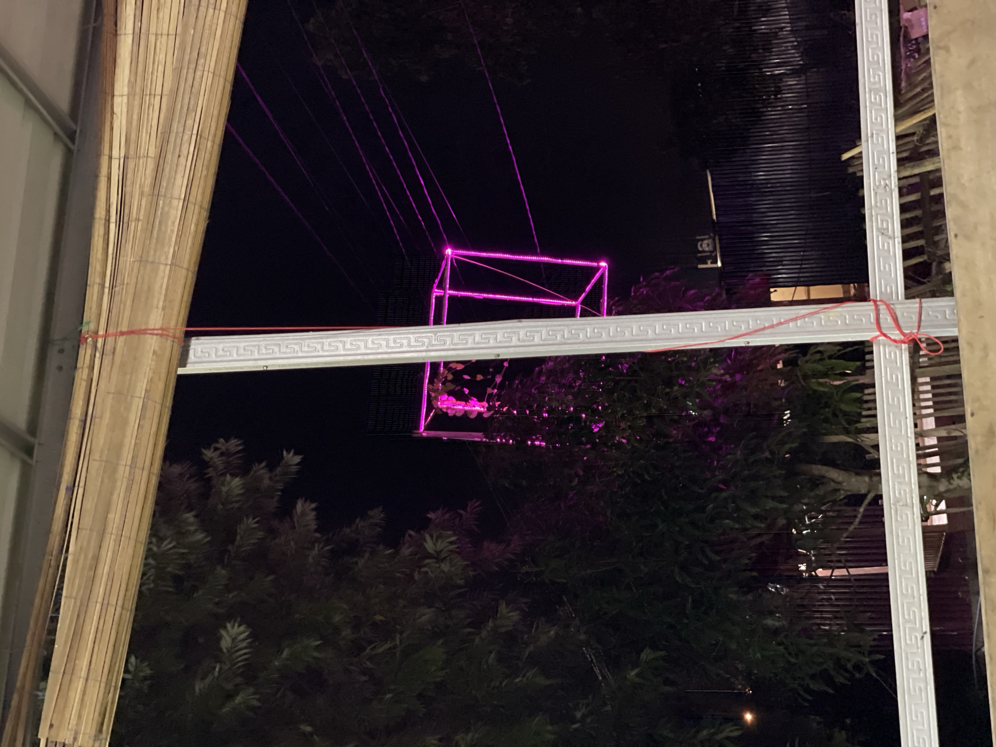 An outdoor night scene in Gampongpaya, Indonesia, features a tall, illuminated pink cube structure against a dark sky. The structure is composed of LED lights forming the edges of a cube, suspended by wires. In the foreground, a bamboo blind hangs from a metal frame, partially obscuring the view. Trees with dark foliage are visible on the left side of the frame, with some branches illuminated by the pink light of the cube. A dark metal fence runs along the bottom of the image. The overall atmosphere is one of urban nighttime illumination and natural elements.