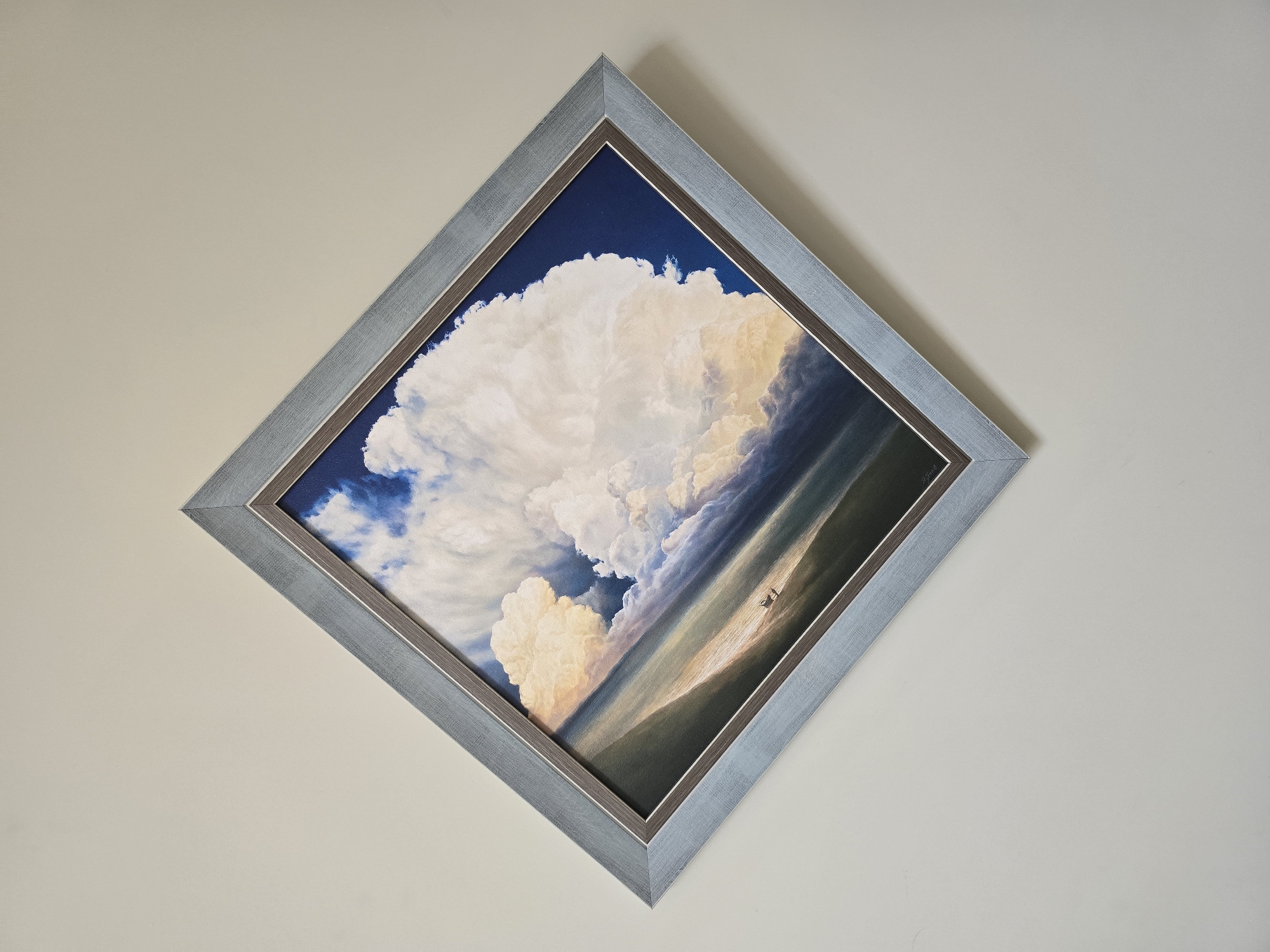 A framed painting, oriented diagonally, hangs on a plain, light beige wall. The artwork depicts a dramatic and expansive sky filled with large, towering white cumulus clouds, likely cumulonimbus formations. The sky transitions from a deep, dark blue at the top left to lighter blues and areas illuminated by what appears to be low-angle sunlight, casting brilliant white and golden highlights on the cloud tops and deep grey-purple shadows within their masses. This lighting suggests either a late afternoon or early morning setting.
Below the prominent cloud formations, a vast, undulating landscape or body of water stretches into the distance, rendered in muted shades of dark green, brown, and ochre, with patches of warm light. Two small, indistinct figures are visible in the mid-ground, appearing to walk or stand on a ridge or distant slope, emphasizing the immense scale of the natural environment and the grandeur of the sky above them.
The painting is encased in a multi-layered frame. The outermost layer is a textured, silvery-grey material with a slightly distressed or weathered appearance, possibly wood. This is followed by a thin, dark brown or bronze inner border before the canvas begins. An illegible signature is faintly visible in the bottom right corner of the painting. The plain, light-colored wall serves as a neutral background, allowing the vibrant and dynamic artwork to be the central focus. The diagonal orientation of the square frame adds a unique visual dynamism to the display.