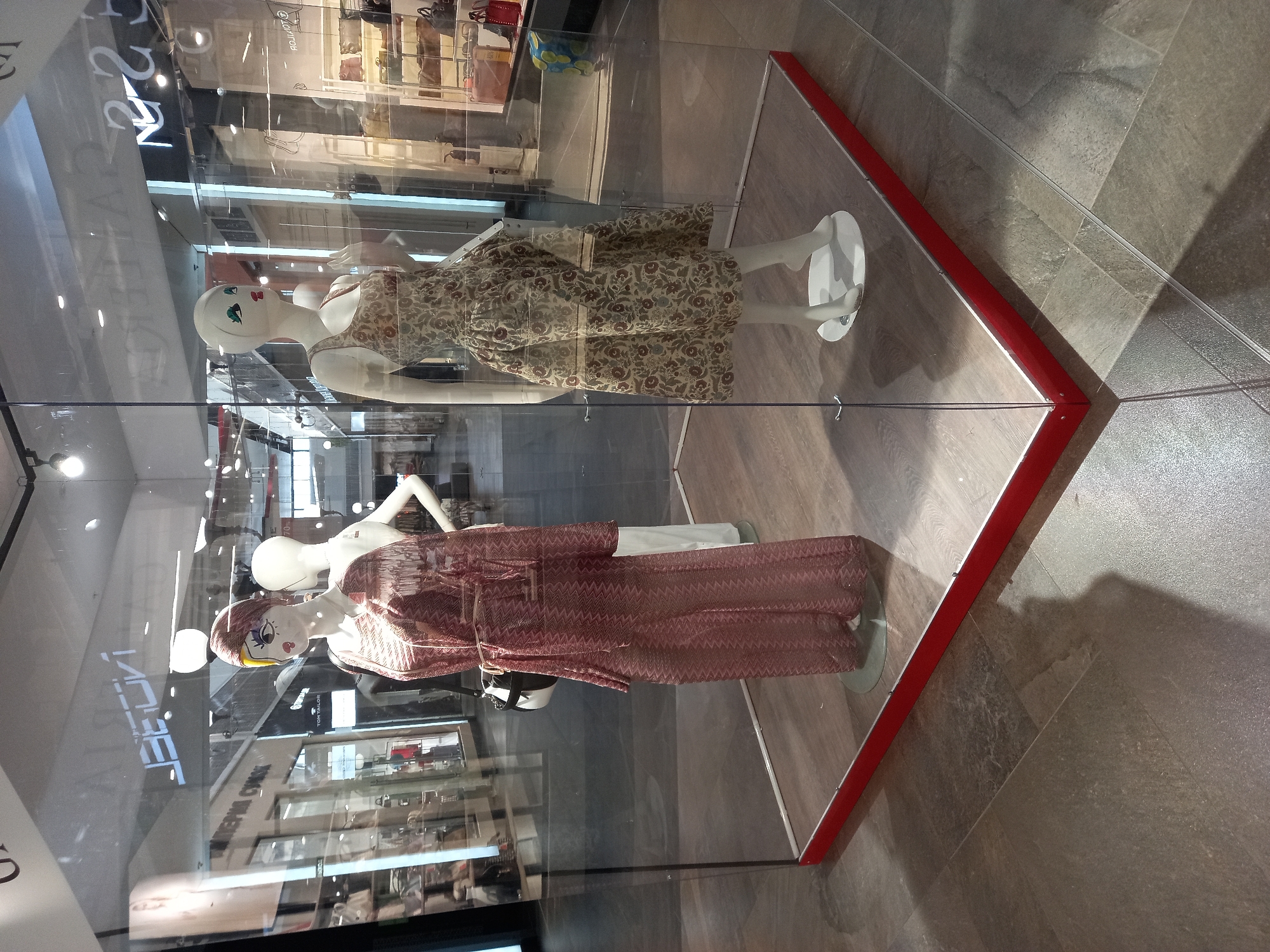 In a shopping mall in Yekaterinburg, Russia, a glass display case features two mannequins. The mannequin on the left wears a long, loose-fitting, patterned pink jumpsuit. It also has a black and white bag slung over its shoulder. The mannequin on the right is adorned in a vintage-style dress with a floral pattern of brown and green on a light beige background. The display case has a red trim at its base. The floor is tiled with a dark, stone-like pattern. The background of the image shows other shops and escalators within the mall.