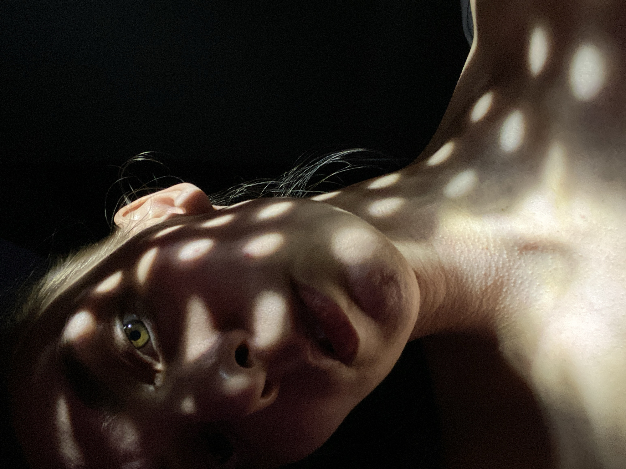 A close-up, low-angle shot depicts a person's face and neck against a dark background. The subject's skin is illuminated by dappled light, creating a pattern of light and shadow across their face and upper chest. The subject's eye is visible. No evidence of the location being Chaikovsky, Russia is present in the image.