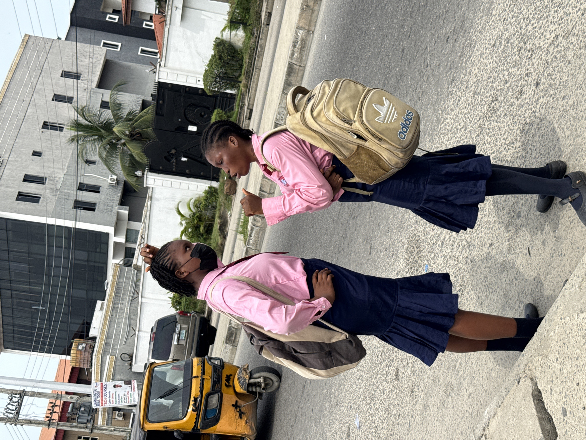 Two young girls in pink long-sleeve shirts, navy-blue skirts, and backpacks stand on a paved surface. The girl on the left wears a black mask. They appear to be in conversation. A yellow three-wheeled vehicle, likely a "keke," is parked nearby. Buildings, including a modern multi-story structure, are visible in the background. A sign is affixed to a utility pole. Based on the reference, this scene takes place in Ado, Nigeria.
