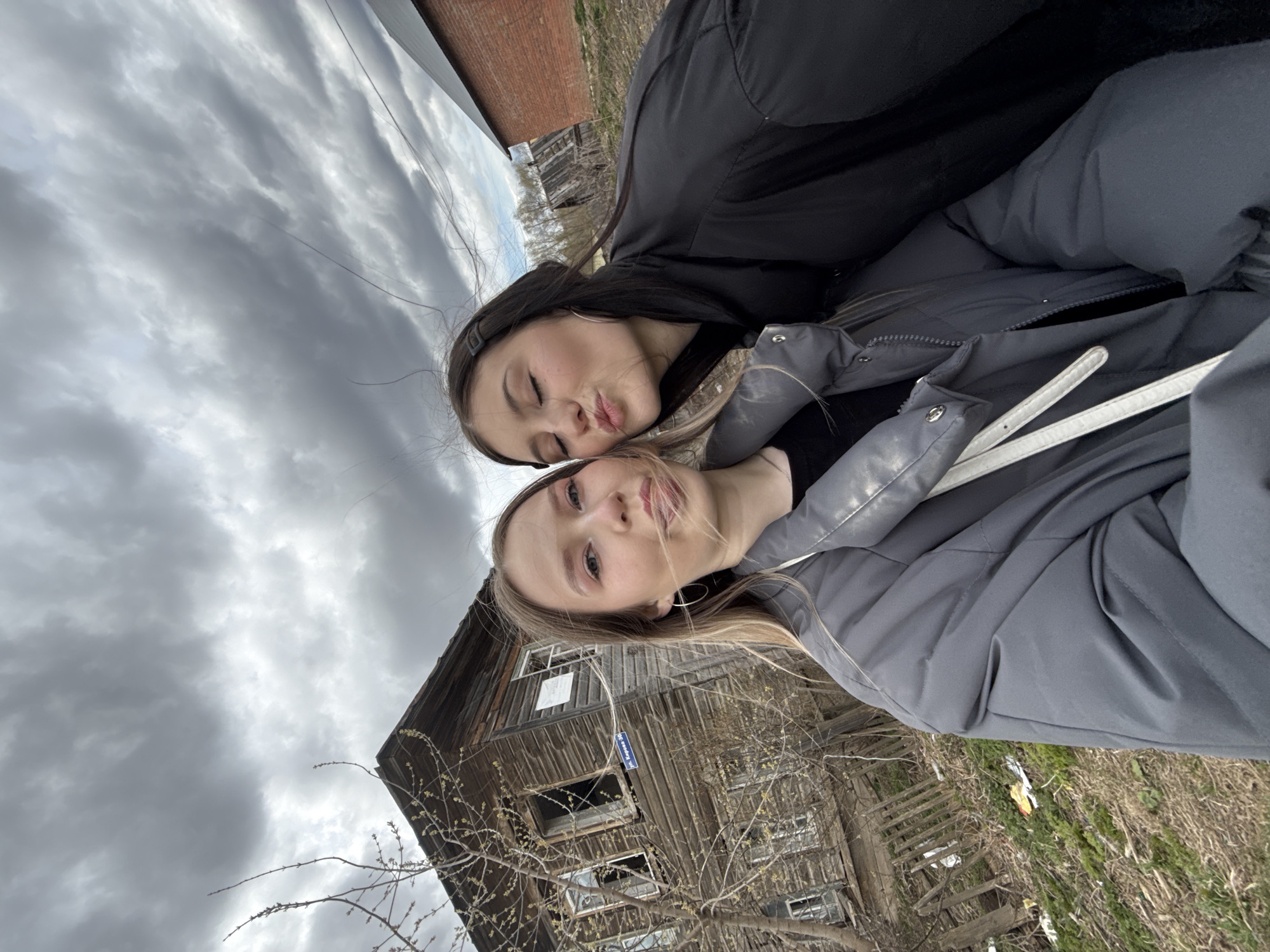 Two young women are featured in a playful selfie, captured outdoors in Foki, Russia. The image is taken from a low angle, looking upwards towards the subjects and the sky. In the foreground, a blonde-haired woman in a grey puffer jacket looks towards the camera with a subtle smile. Behind and slightly above her, a dark-haired woman in a black jacket is making a kissing or pouting face with her eyes closed. Both appear to be wearing warm attire, suitable for cool weather.
The background consists of an old, weathered wooden building, possibly a house or barn, with several broken window frames. Bare tree branches with visible small buds are in front of the building, suggesting early spring. On the building, a blue sign with white text clearly reads "ул. Карла Маркса 36," indicating "Karl Marx Street 36." The ground below is covered in dry grass with some scattered litter. The sky is heavily overcast with prominent grey clouds, creating soft, diffused lighting. The scene conveys a casual and lighthearted moment between the two individuals.
