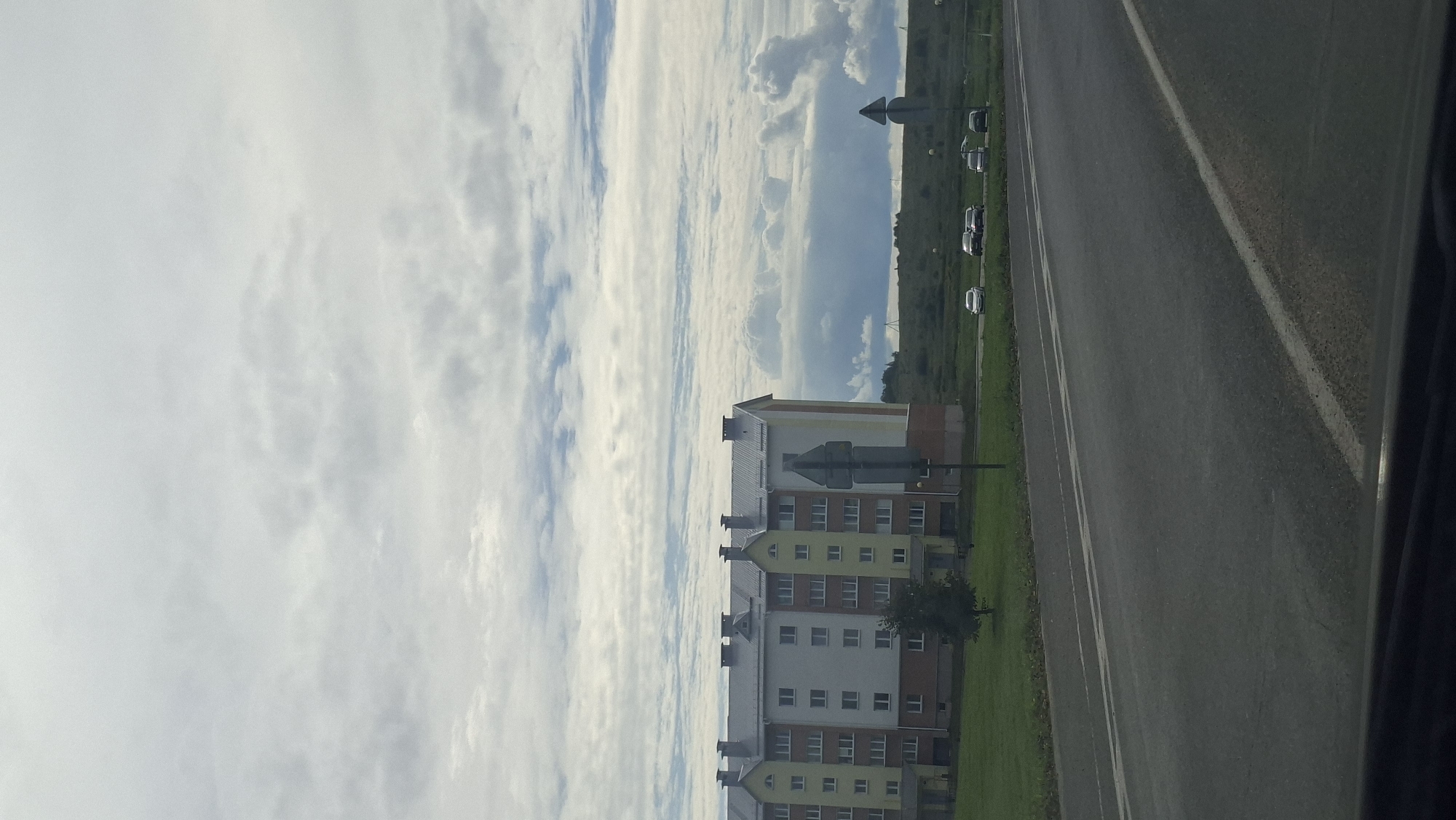 An overcast sky with wispy clouds dominates the upper half of the image. Below, a long, multi-story apartment building with a light yellow and white facade and a grey roof is visible. The building has numerous windows and decorative gables. In front of the building is a grassy area with a single tree. A road runs horizontally across the foreground, with two lanes indicated by white lines. Several cars are parked on the side of the road or driving past the building. Two traffic signs, one a diamond shape with a horizontal line and the other a triangle with a horizontal line, are present on the right side of the road. The scene appears to be on the outskirts of Myadzyel, Belarus, characterized by a mix of residential buildings and open, rural-like terrain.