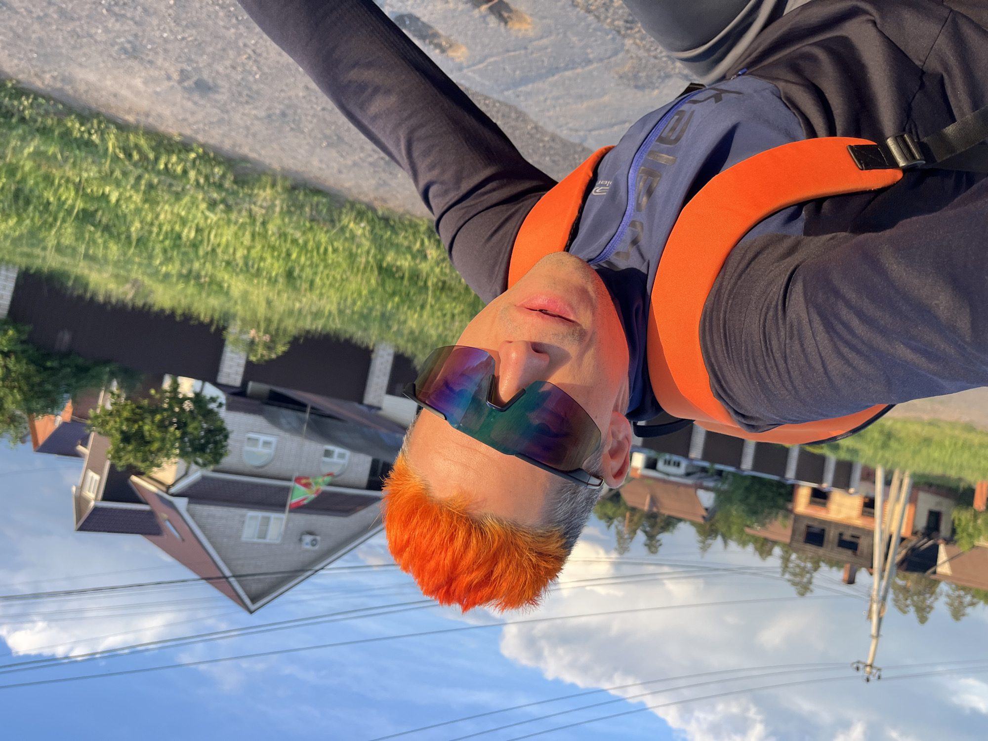 A man with orange hair is wearing reflective sunglasses and a backpack with orange straps. He is standing on a paved surface in the city of Chaikovsky, Russia. In the background are houses, greenery, and a cloudy sky. A flag is visible on one of the houses.